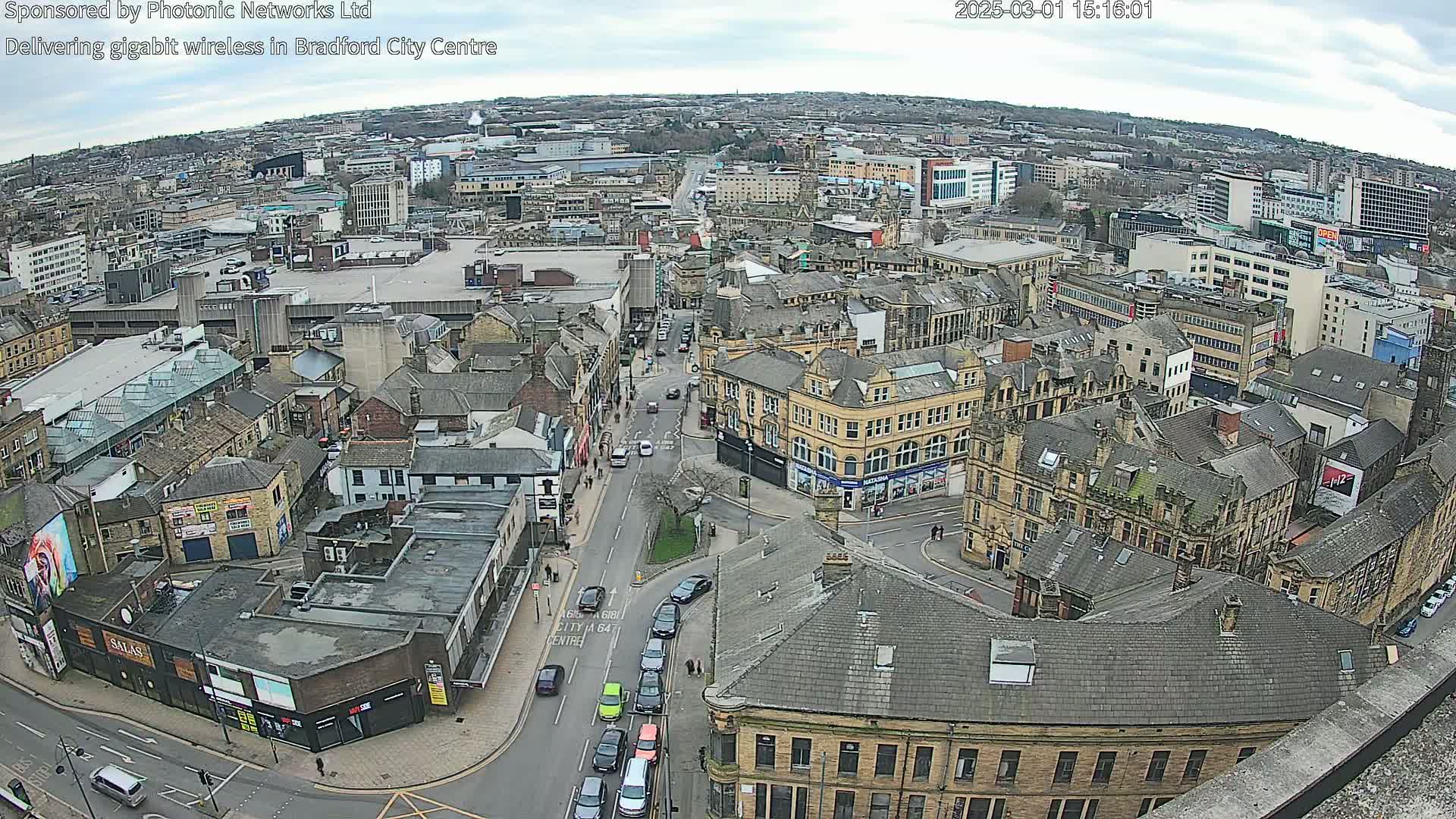 An aerial view of a city center on an overcast day shows a network of streets and buildings, with several cars visible on the roads.
