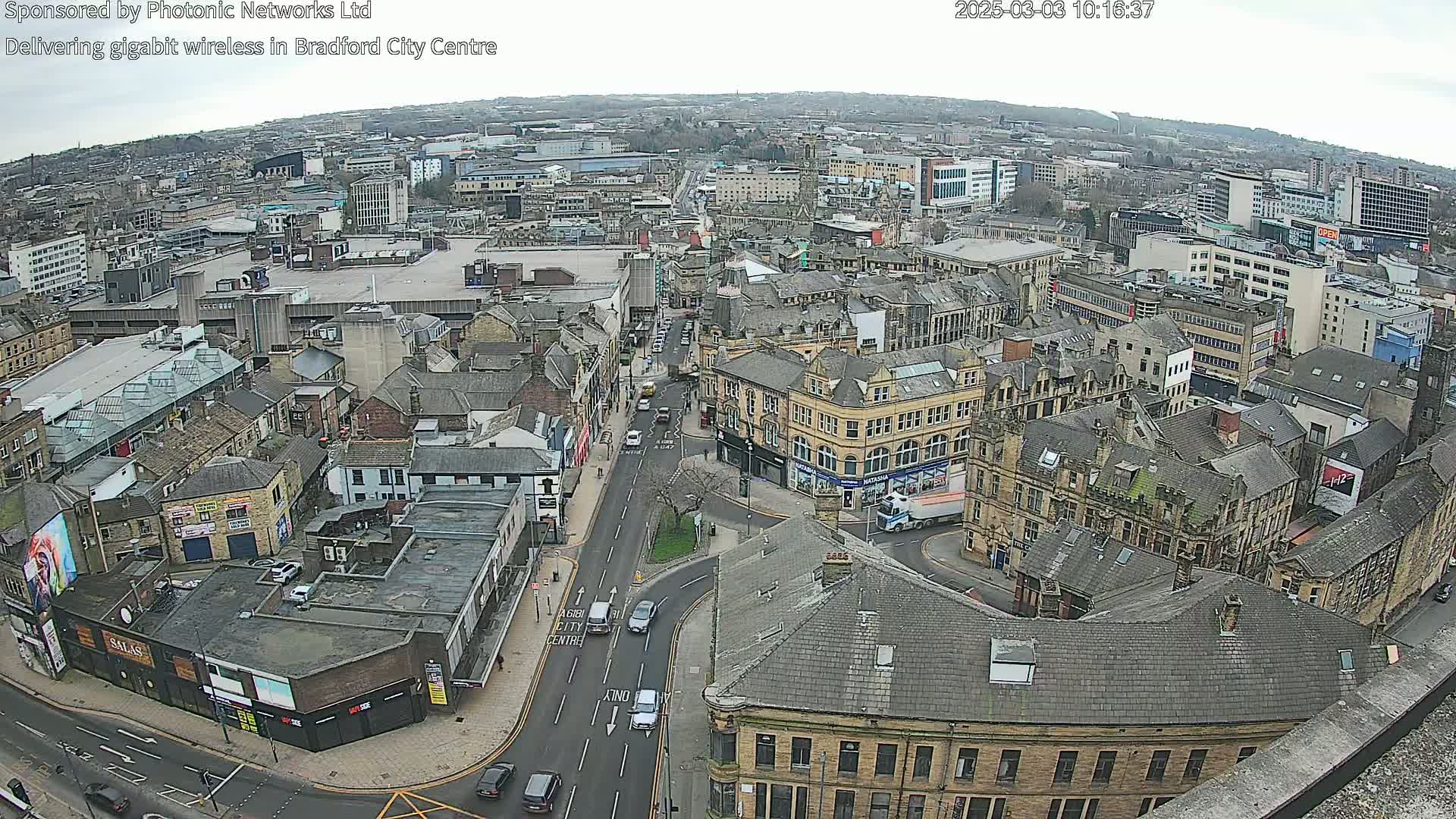 A high-angle, long shot of a city center on an overcast day shows a street intersection with several cars and buildings of varying heights and architectural styles.