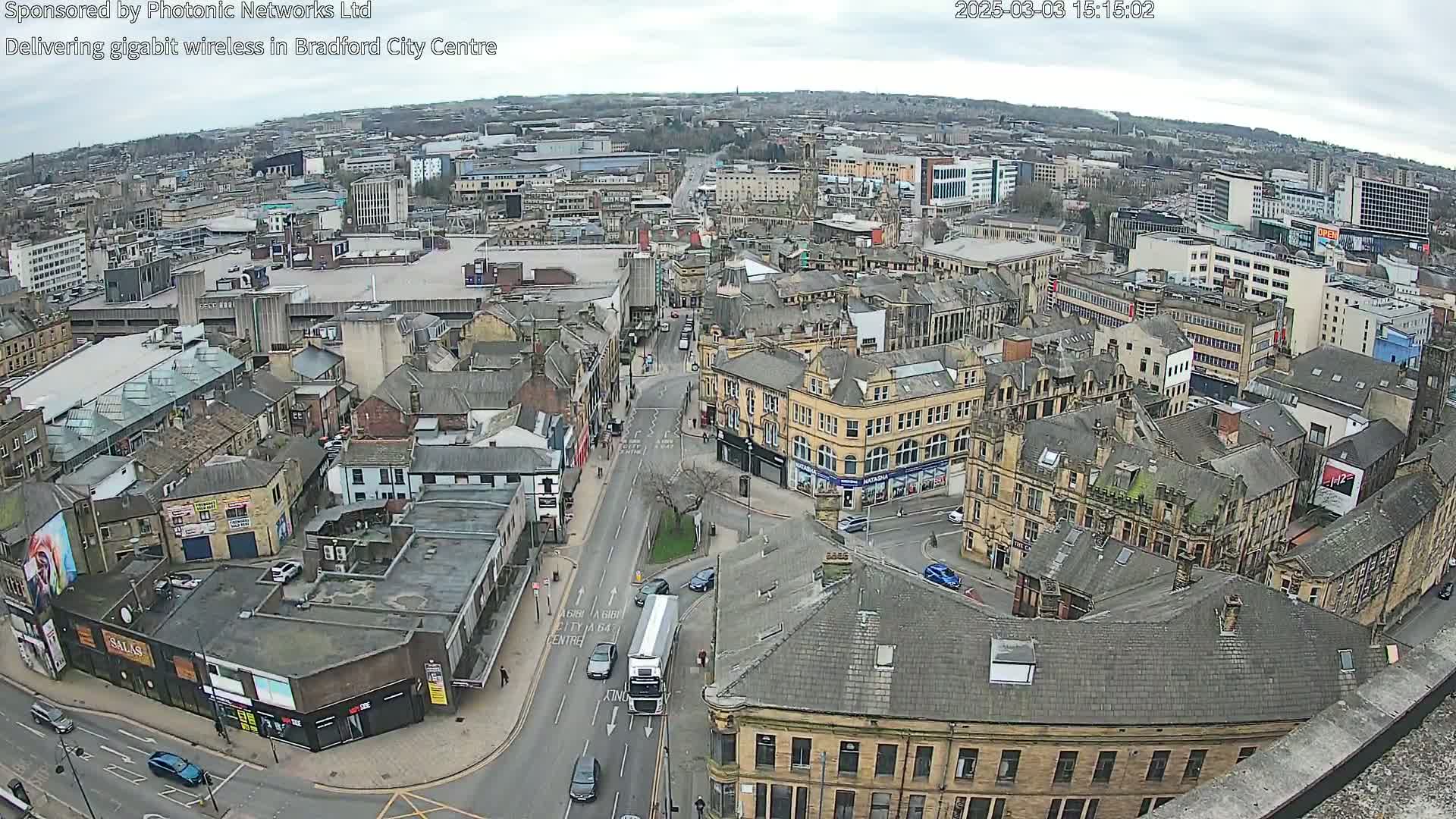 A high-angle, long shot of a city under an overcast sky shows a street scene with several vehicles and many buildings.
