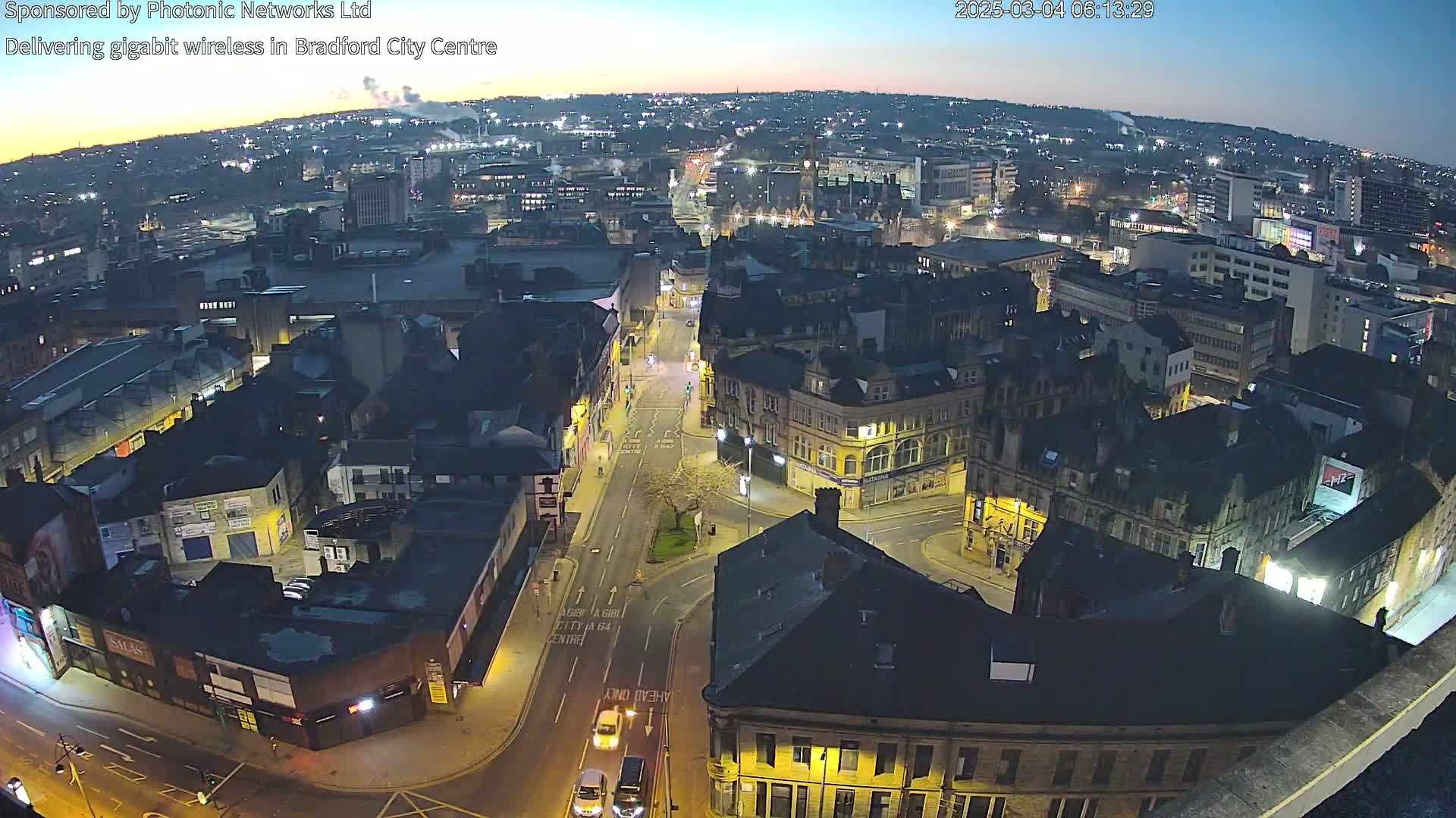 An aerial nighttime view of a city center, showing a network of streets, buildings illuminated by streetlights, and a twilight sky.