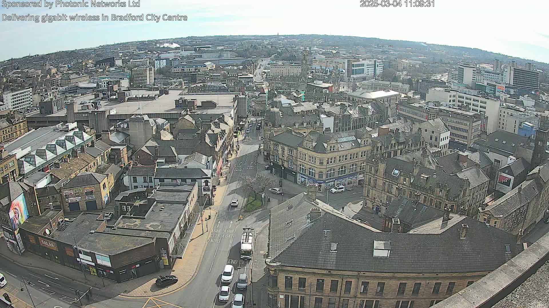 A high-angle, long shot of a city center on a sunny day, showing numerous buildings, streets, and a few vehicles.