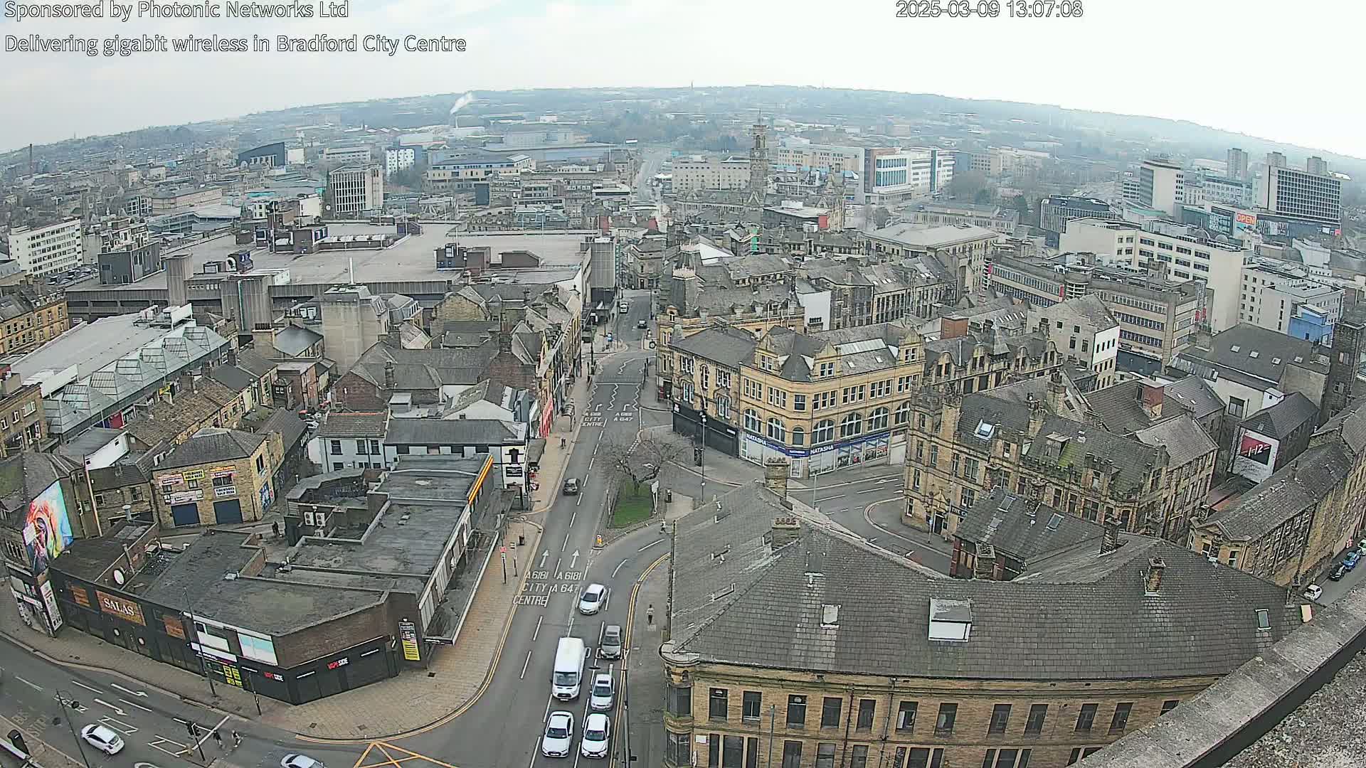 An aerial view of a city on an overcast day shows several streets intersecting, with cars driving on them and many buildings of varying heights and architectural styles.