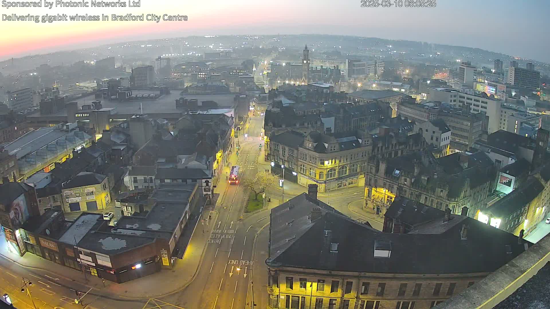 A hazy, twilight view of a city center with illuminated buildings and streets, showing a bus on a road at a crossroads.