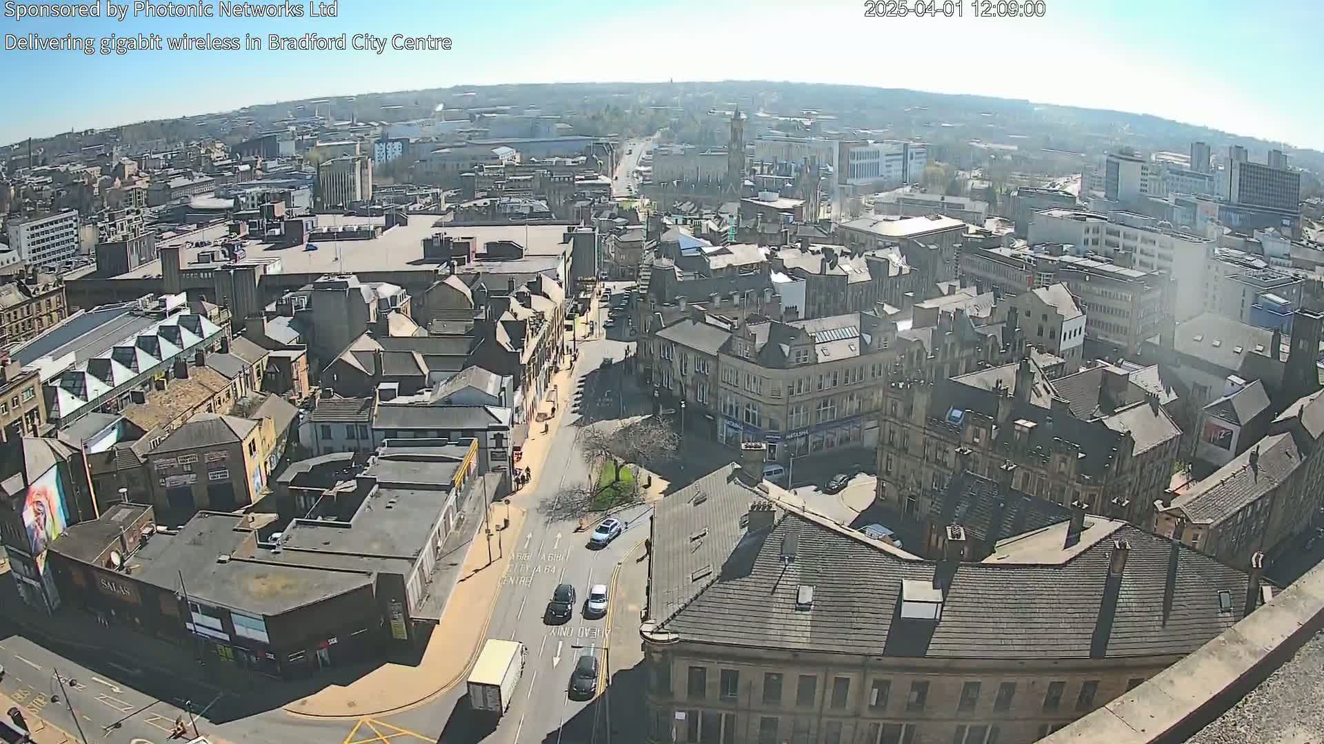 A high-angle, sunny view shows a city center with many buildings, streets, and a few cars.