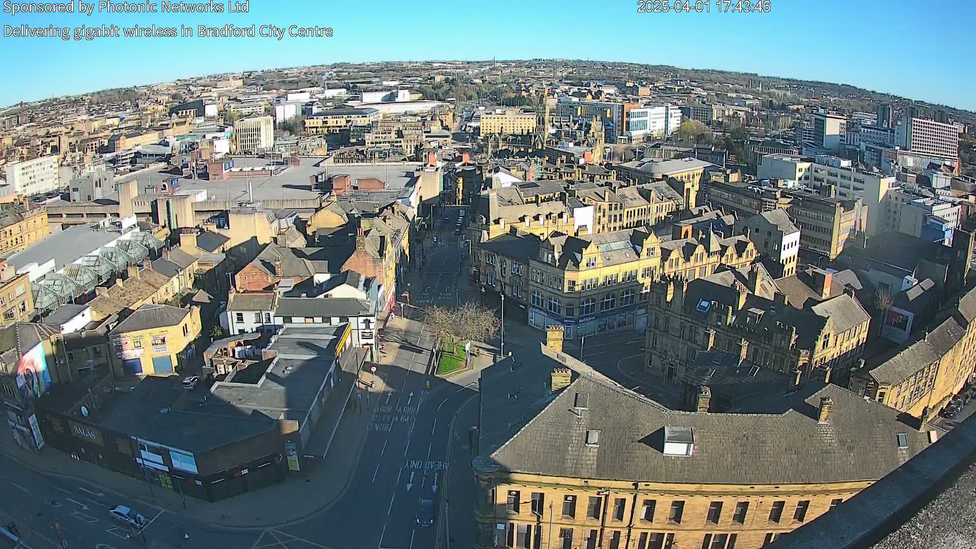 A high-angle, sunny view of a city center with numerous buildings, streets, and a large parking area.