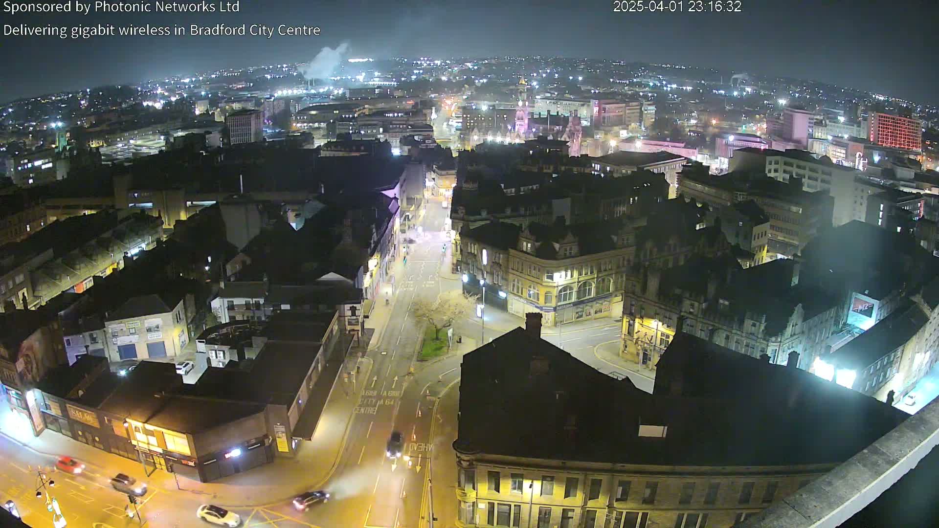 A nighttime, high-angle view of a city center with illuminated buildings and streets, showing sparse vehicle traffic.