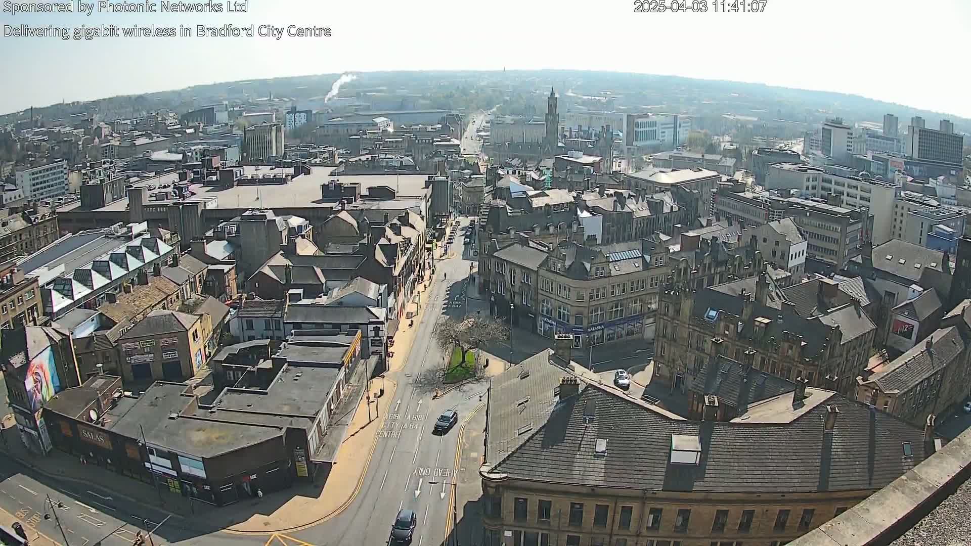 A high-angle, sunny-day view of a city center with numerous buildings and a street intersection, showing vehicles and some pedestrian activity.