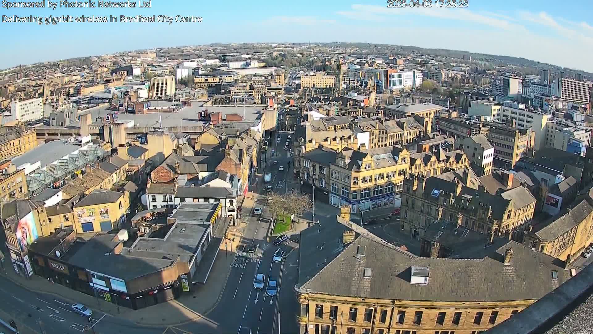 A high-angle, sunny view of a city center with many closely packed buildings and streets, showing vehicles and pedestrians.
