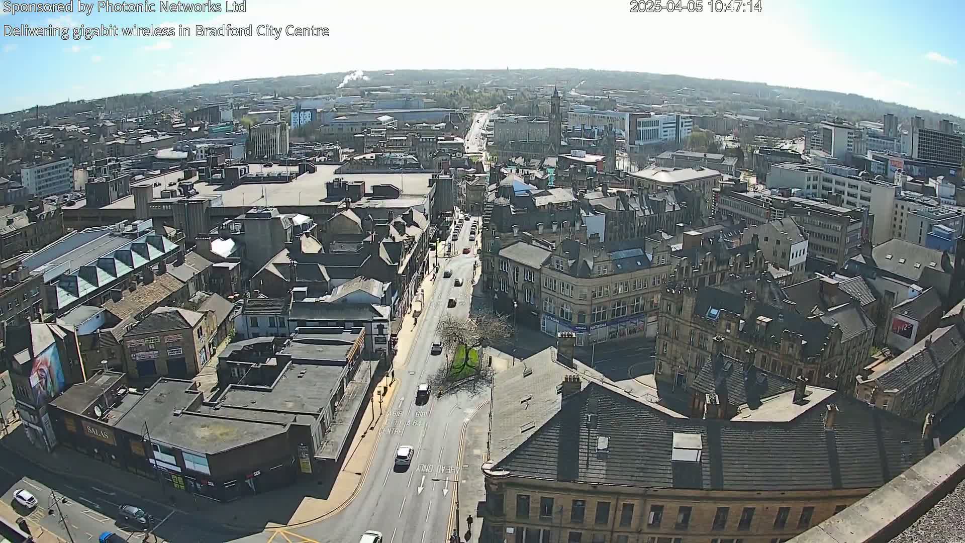 A high-angle, long shot on a sunny day shows a city center with many buildings and a road with cars.