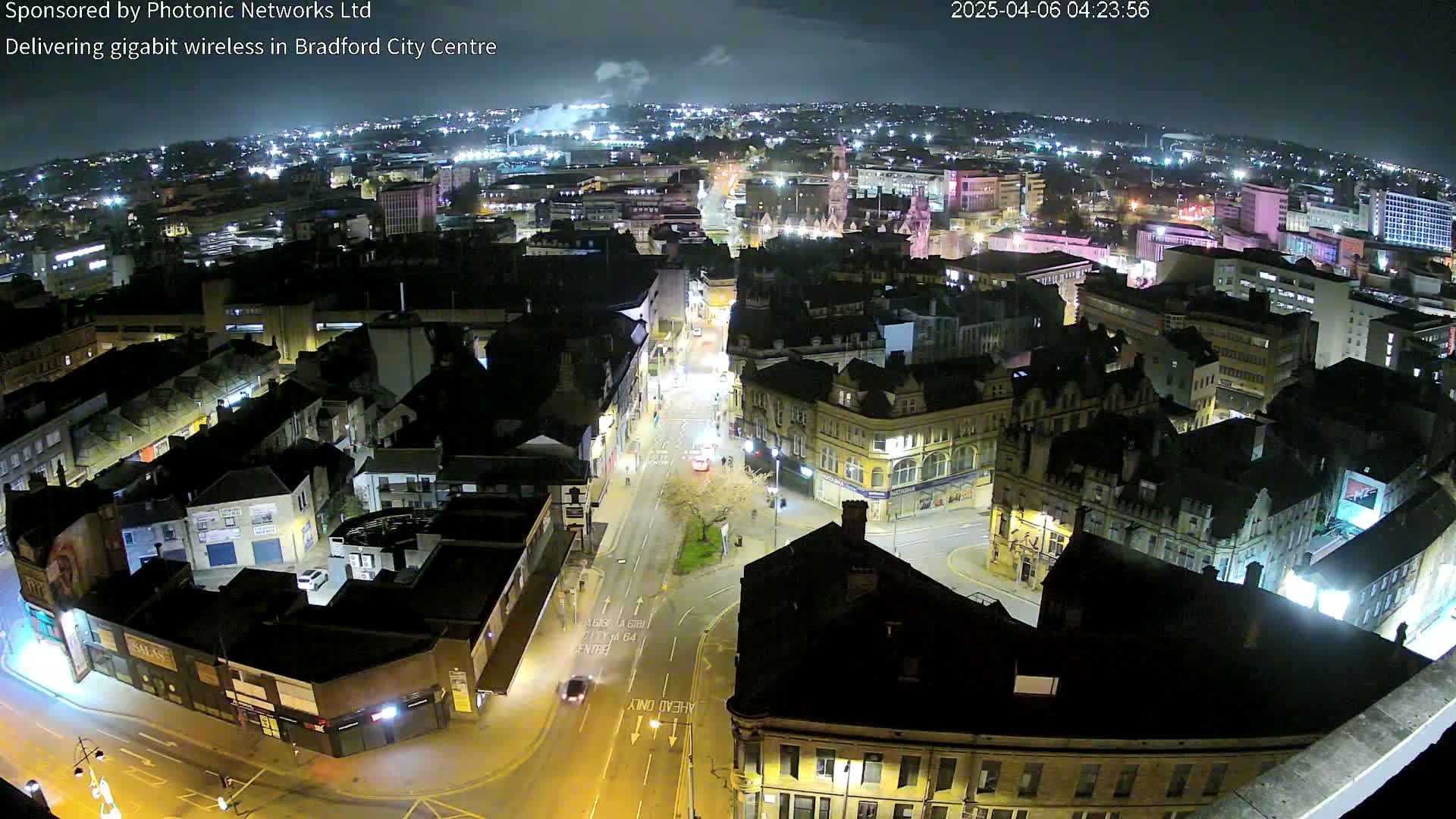 A nighttime aerial view of a city center, showing illuminated buildings and streets with minimal traffic under a partly cloudy sky.