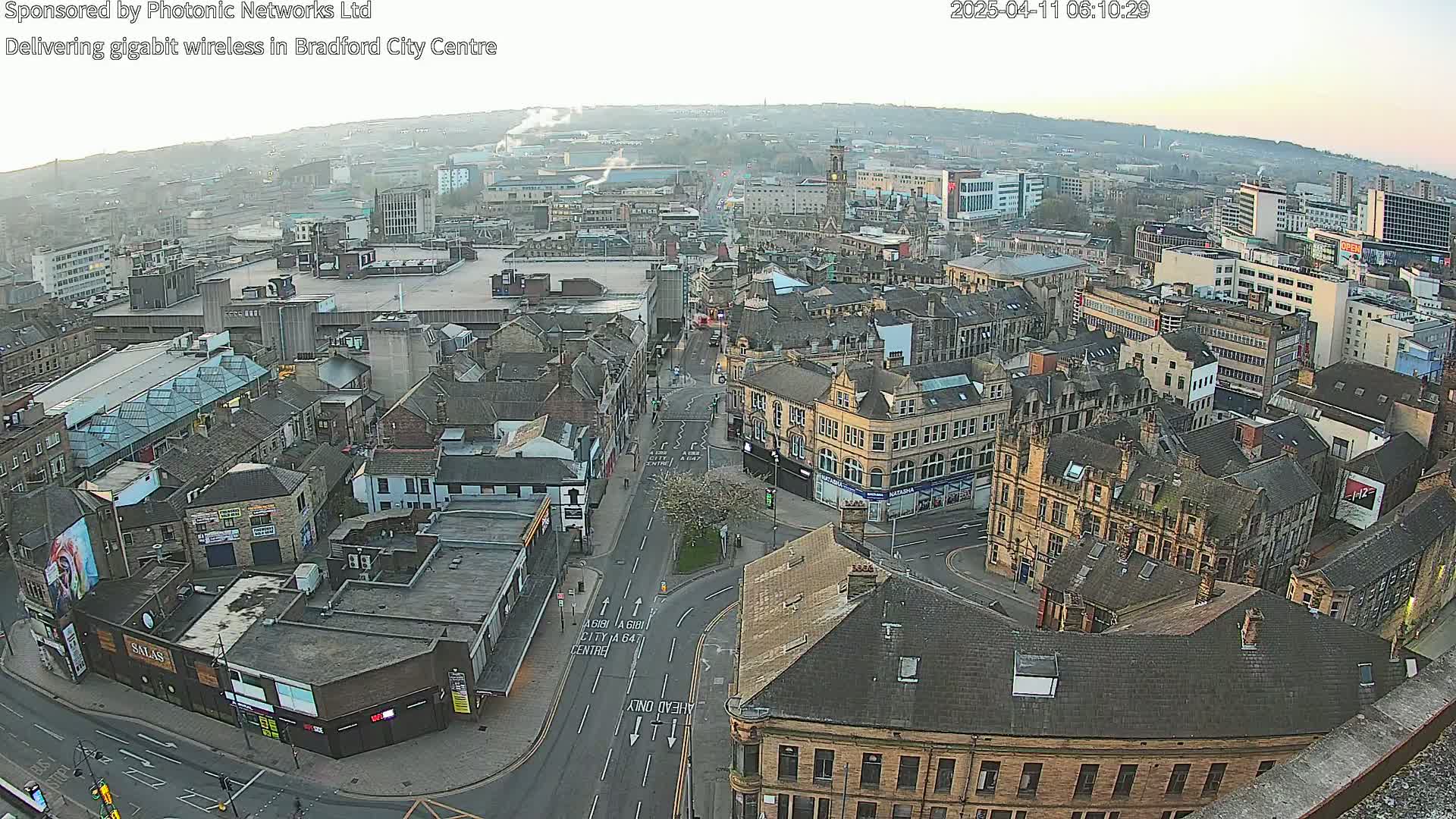 A high-angle, long shot of a city center on a hazy day shows many buildings of various sizes and styles, with streets and roads weaving between them.