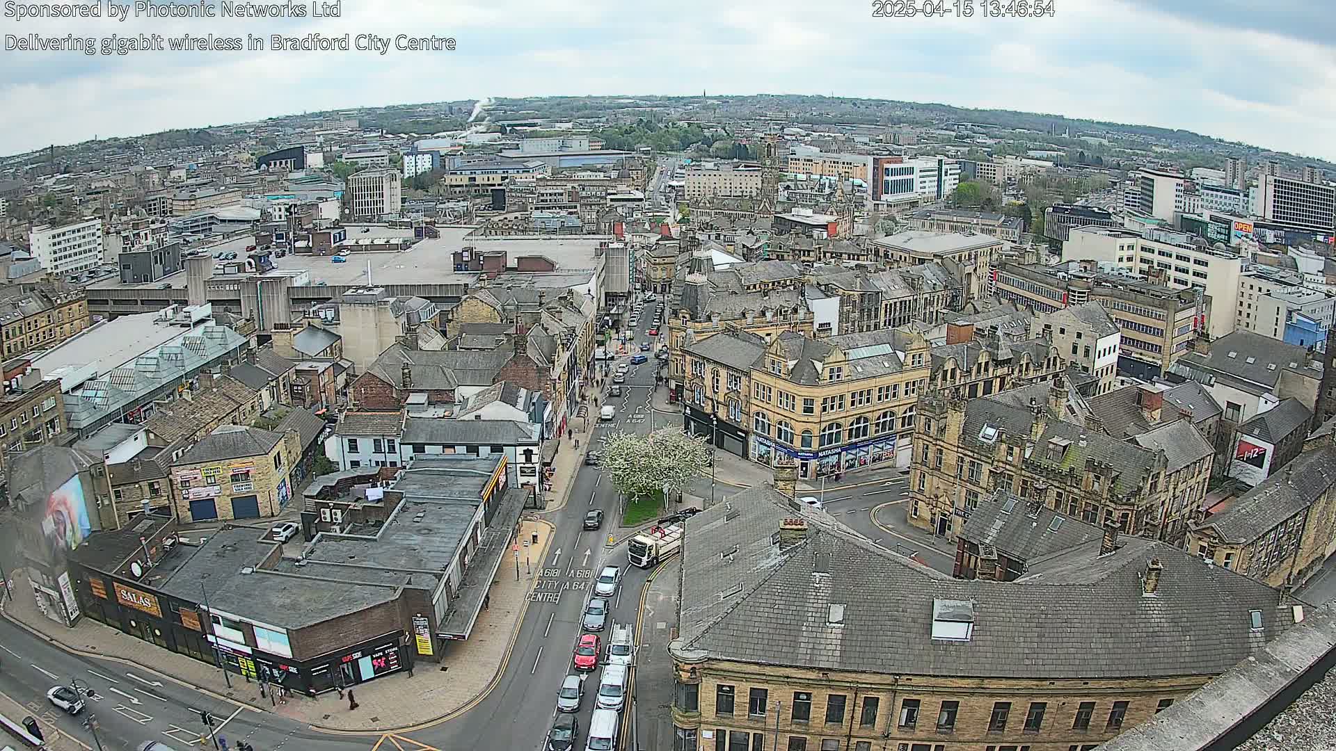 A high-angle, long shot of a city center on a partly cloudy day, showing many buildings and streets with cars and pedestrians.