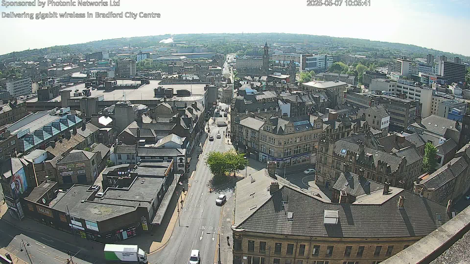 A high-angle, long shot of a city on a sunny day shows many buildings and streets with sparse vehicle traffic.