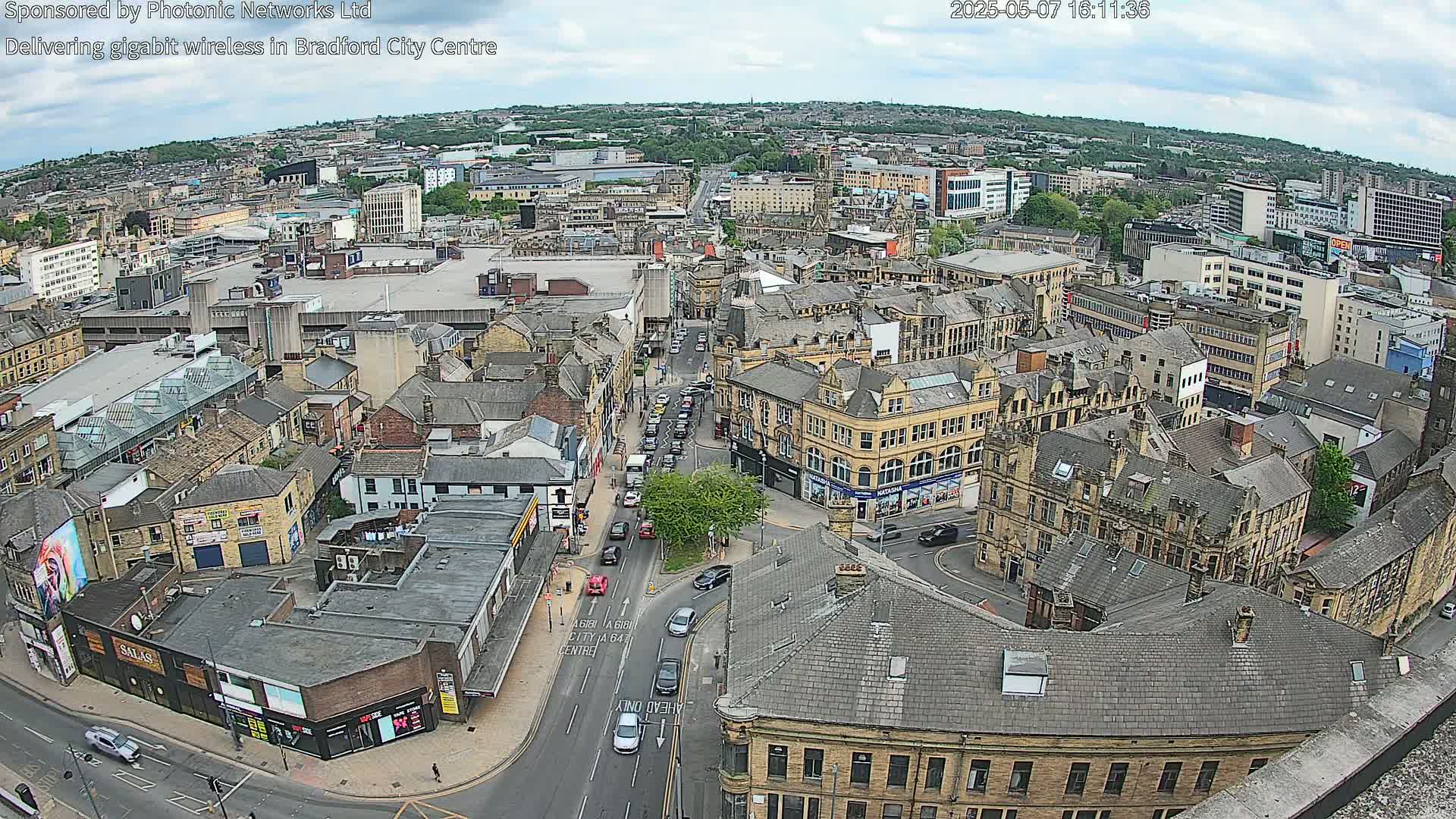 A high-angle, daytime view of a city center with many buildings, roads, and vehicles, under partly cloudy skies.