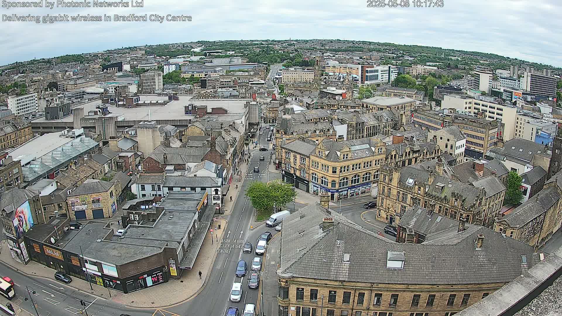An aerial view of a city center on an overcast day, showing numerous buildings and streets with some cars and pedestrians visible.