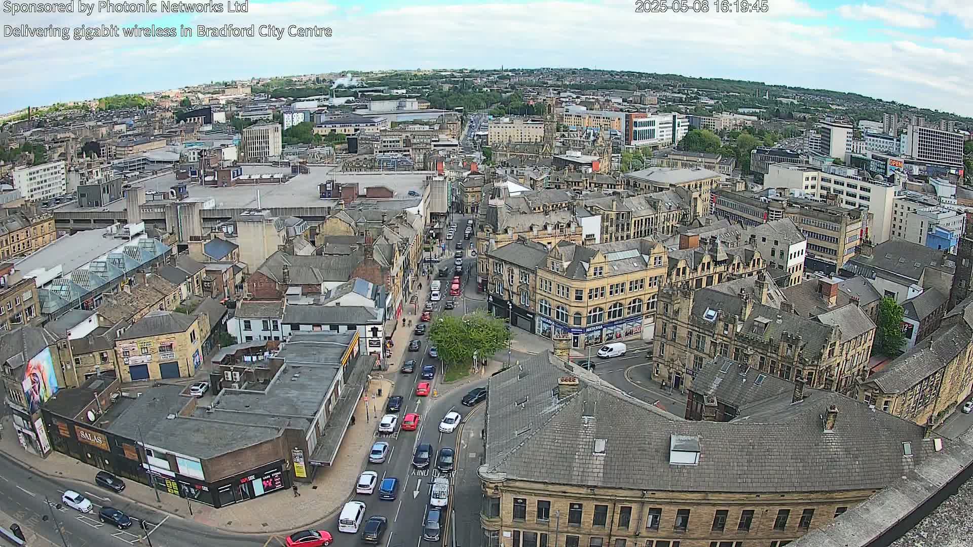 A high-angle view of a city center on a partly cloudy day, showing many buildings and traffic on the streets below.
