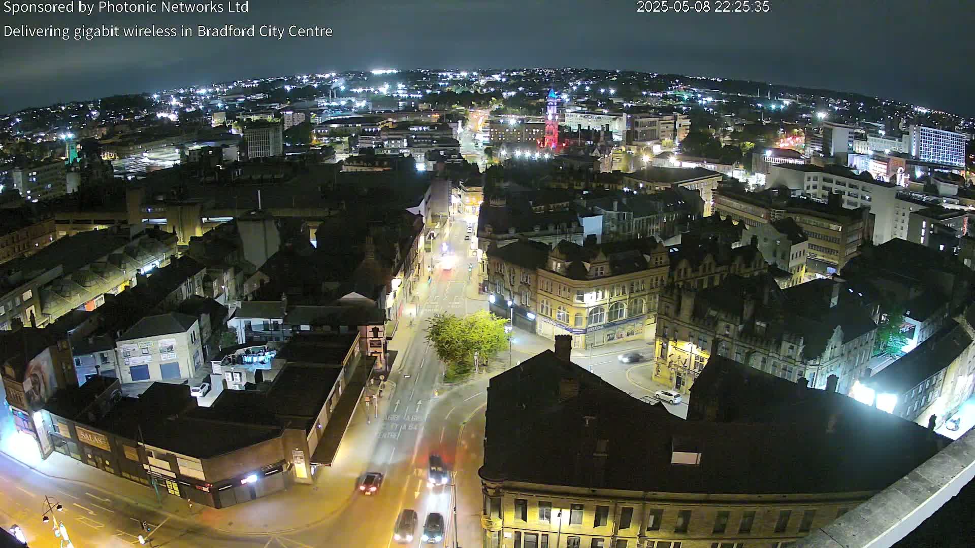 A nighttime, high-angle view of a city center with illuminated buildings and streets, showing several cars driving on roads.