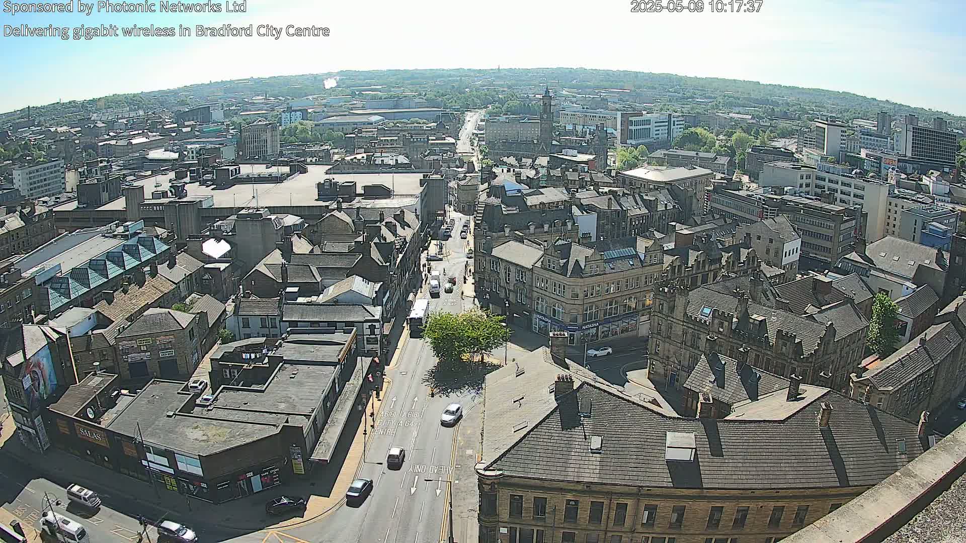 A high-angle, long shot of a city on a sunny day shows many buildings and streets with several cars driving on the roads.