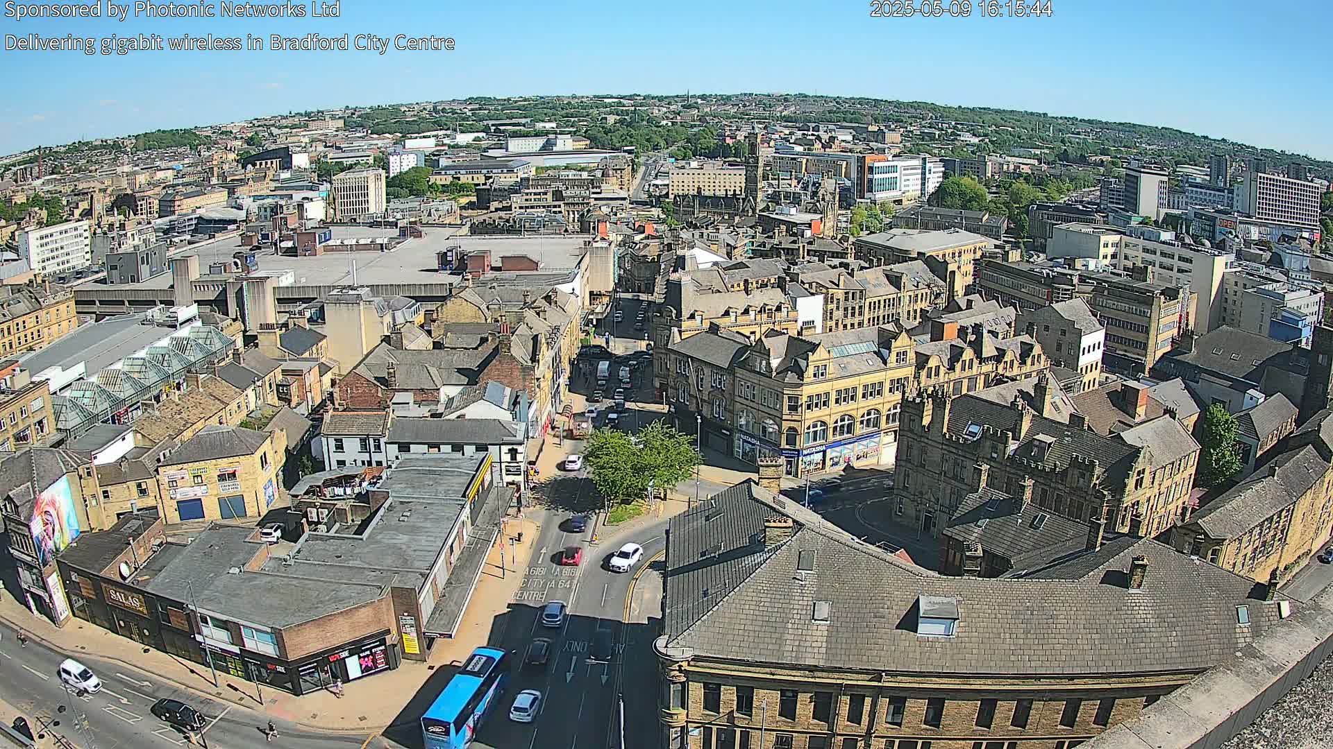 A high-angle, long shot on a sunny day shows a sprawling city center with numerous buildings and streets, featuring vehicles and a bus on the roads.
