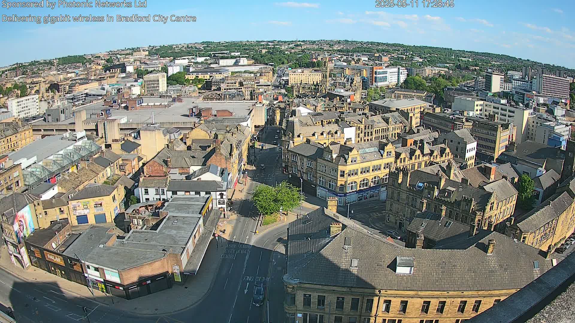 A high-angle, sunny view of a dense cityscape shows many brownstone buildings and streets, with a large parking lot visible in the middle distance.