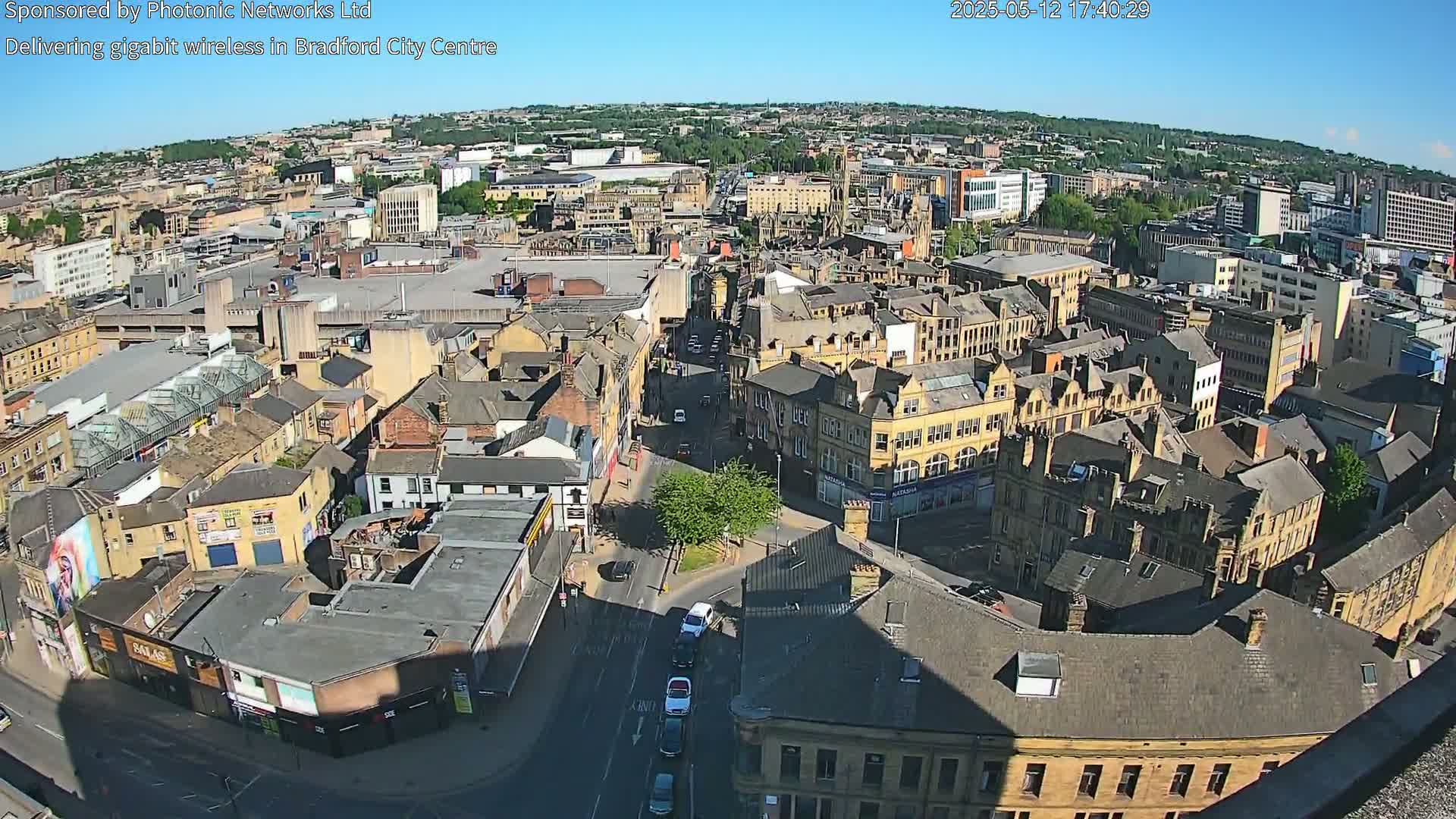 Here is a description of the image:

A high-angle, long shot on a sunny day shows a dense collection of buildings in a city center, with several streets visible and cars driving on them.