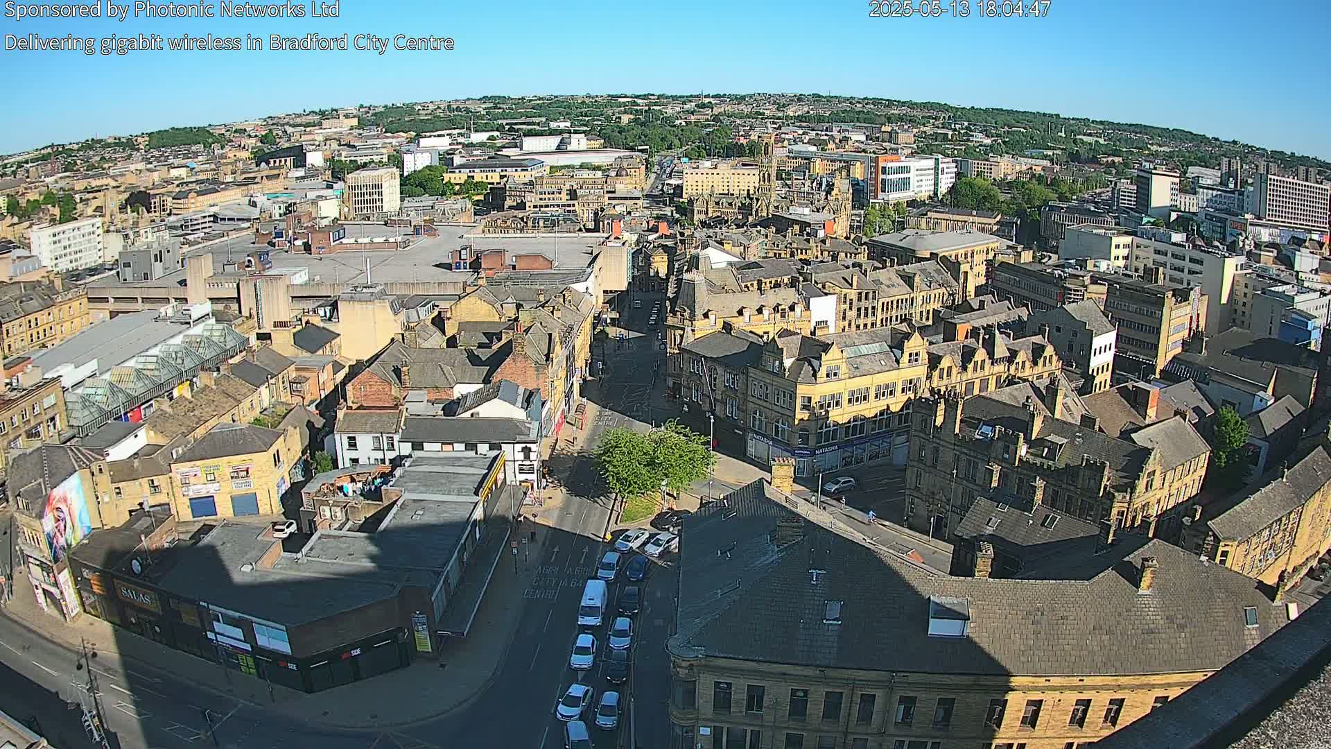 A high-angle, long shot of a city on a sunny day shows numerous buildings and streets, with a few cars visible on a roadway.
