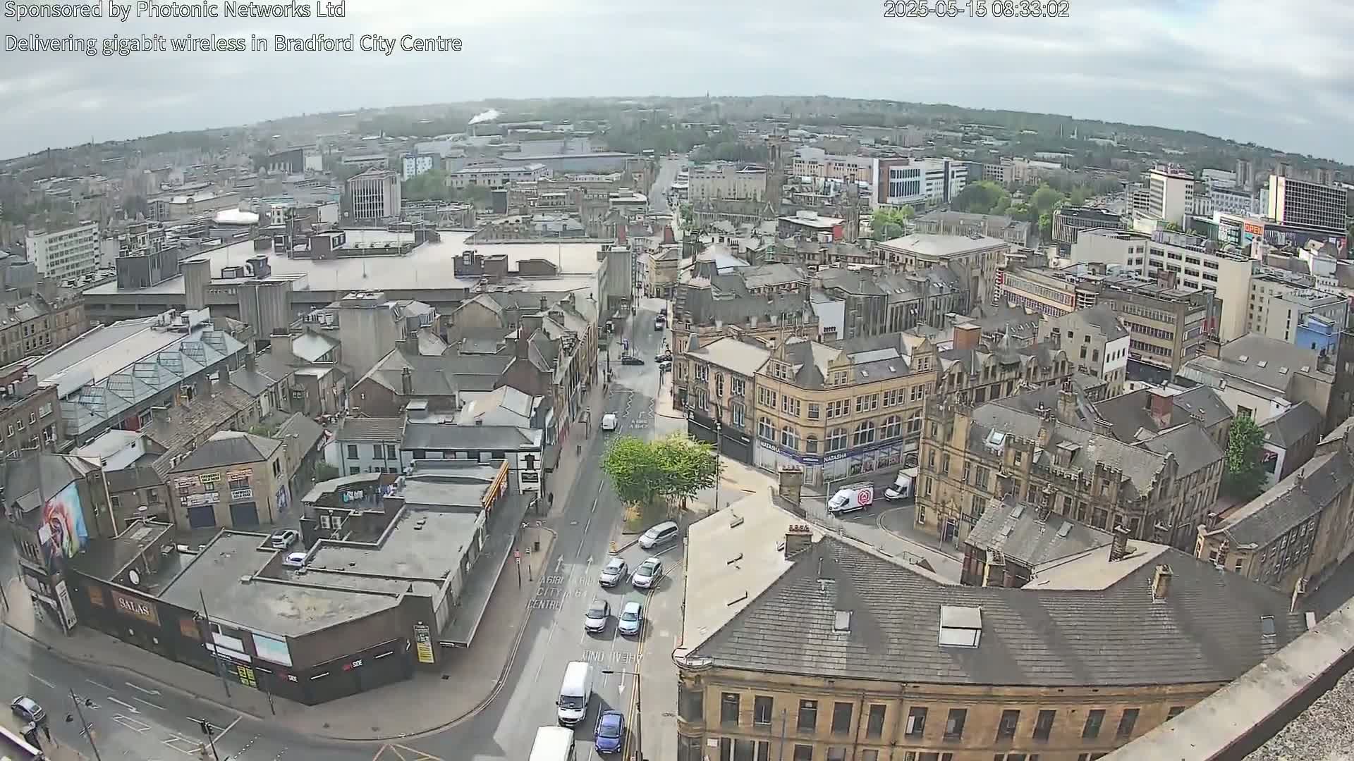 Here is a description of the image:  A high-angle, long shot of a city center on an overcast day shows many buildings and a few cars on the street.