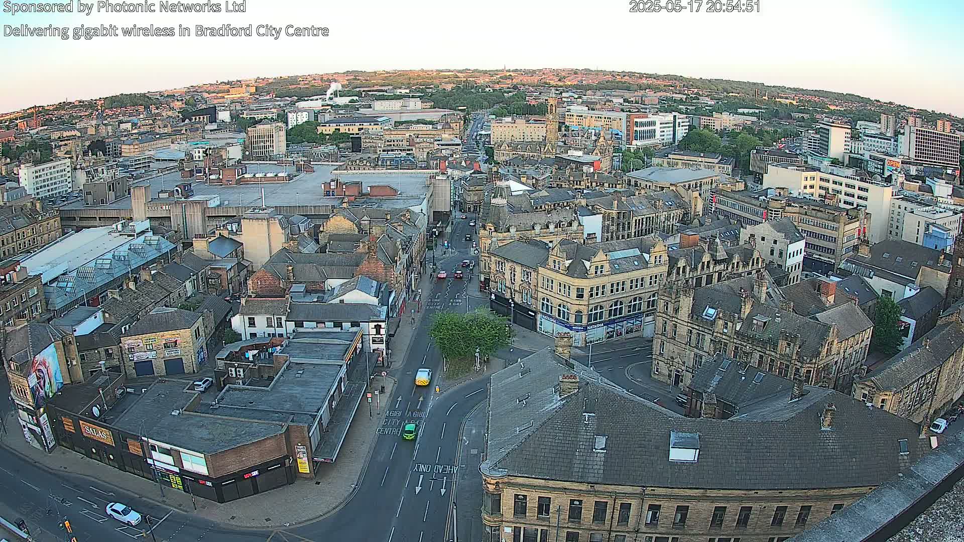 A high-angle, wide shot of a city center at dusk shows many buildings and streets, with a few cars visible on the roads.