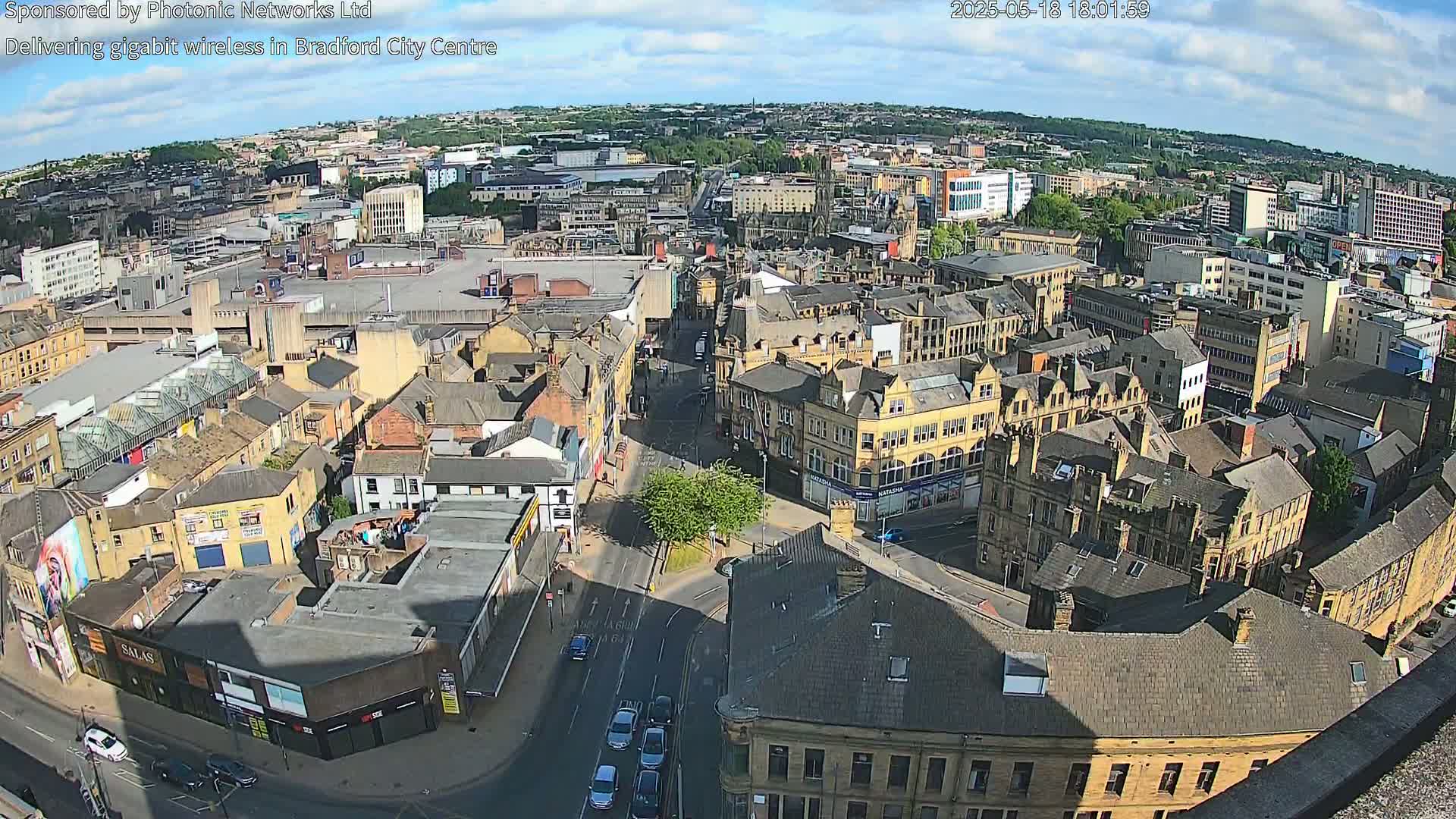 A high-angle, sunny view of a dense city center with numerous buildings, streets, and some vehicles, under partly cloudy skies.