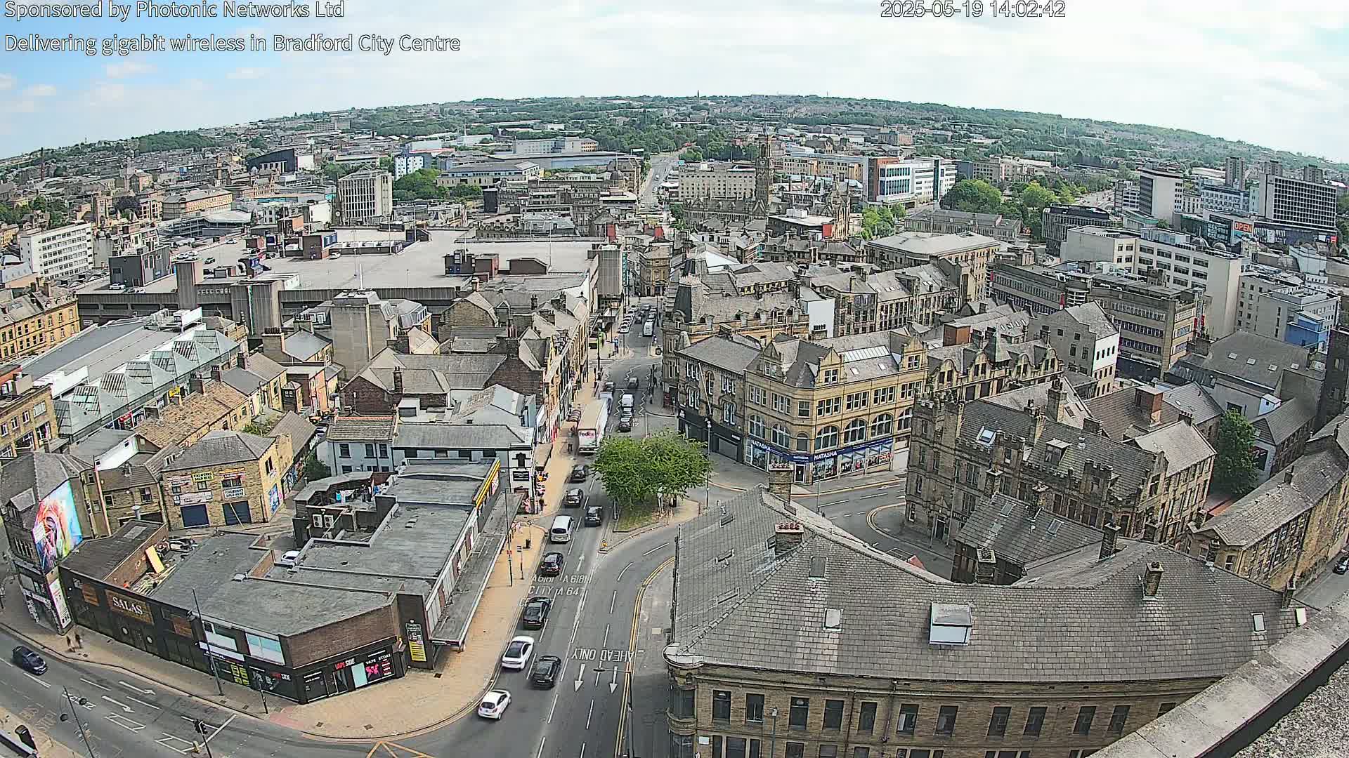 A high-angle, daytime view of a city center with numerous buildings, streets, and vehicles under partly cloudy skies.