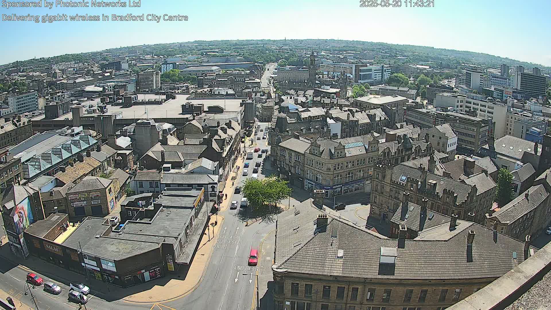 An aerial view of a city on a sunny day shows a cityscape of closely packed buildings, streets with cars, and a large square or plaza in the center.