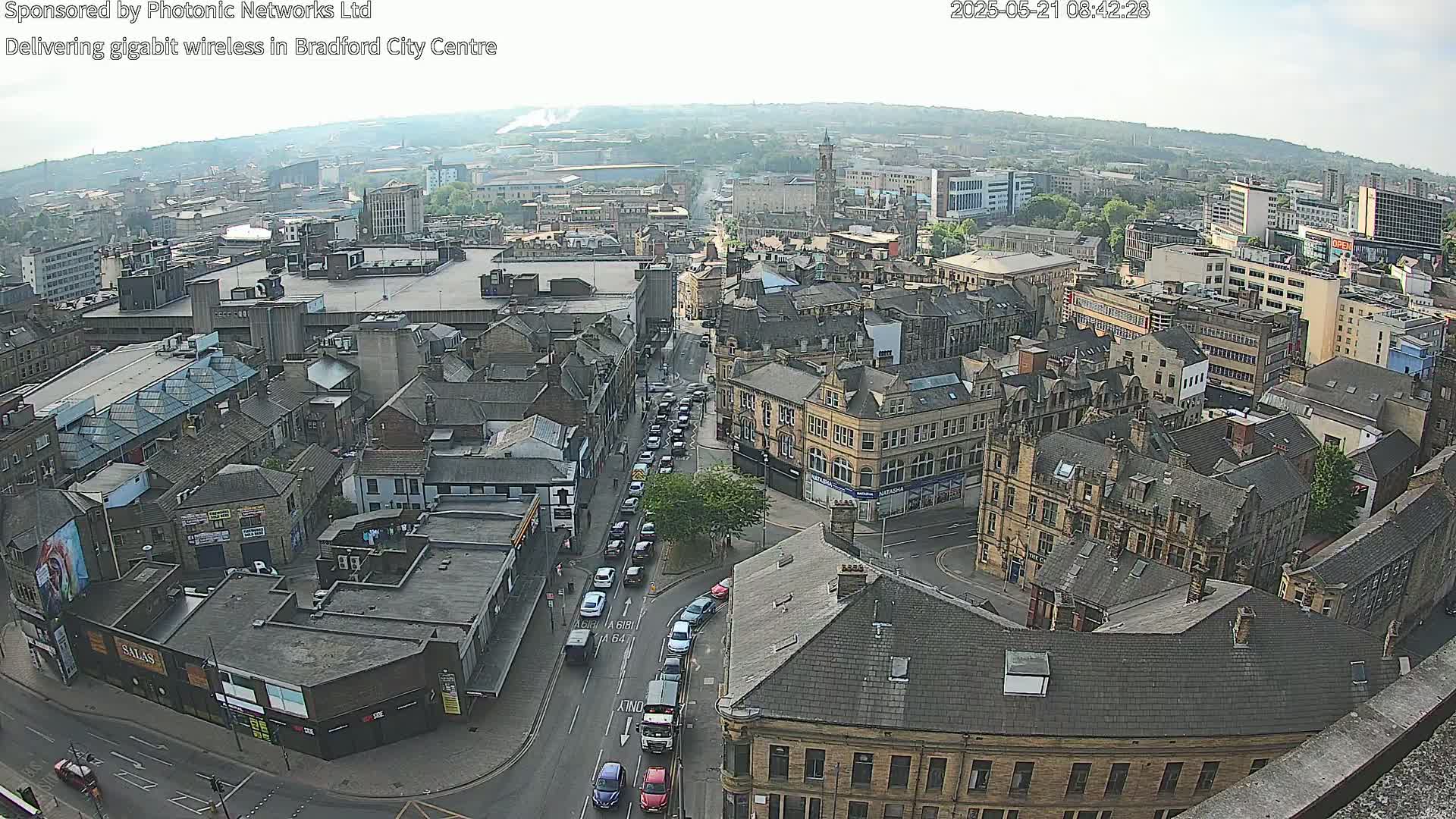 A high-angle, long shot on a partly sunny day shows a city center with many buildings and roads, including traffic on a main road.