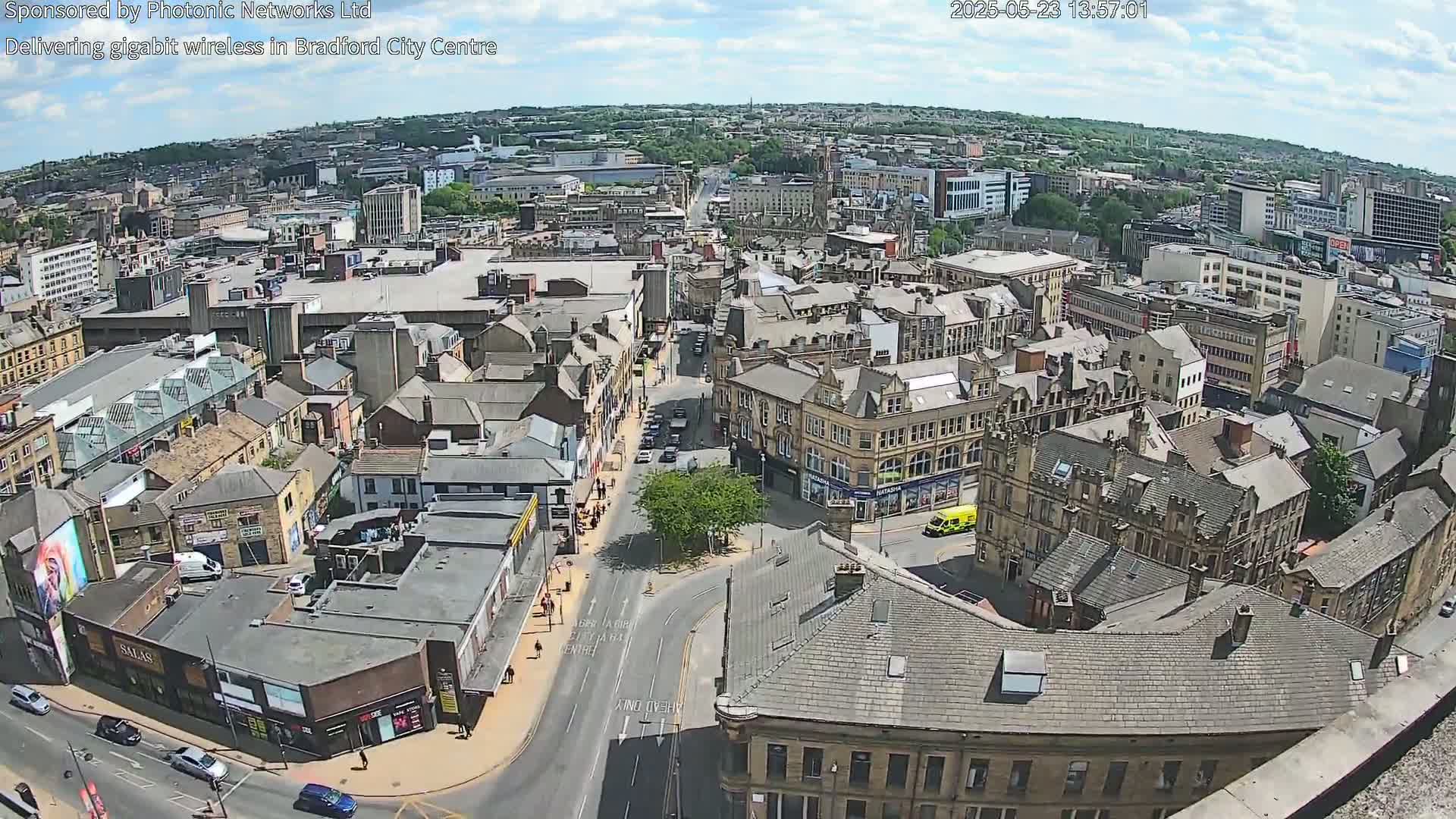 Here is a description of the image: A high-angle, long shot of a city on a partly sunny day shows numerous buildings and streets with some cars visible.