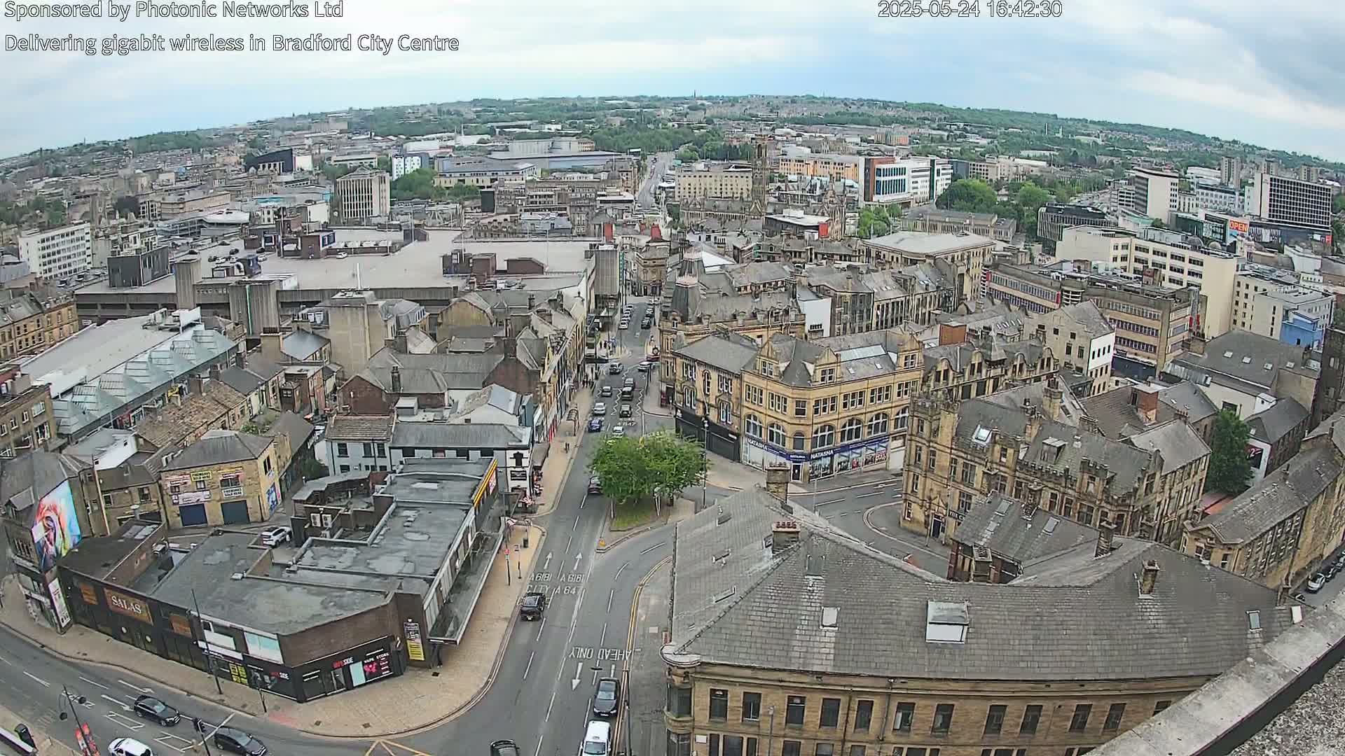 A high-angle, long shot of a city center on an overcast day shows a dense collection of buildings, streets with cars, and a tree in a small roundabout.