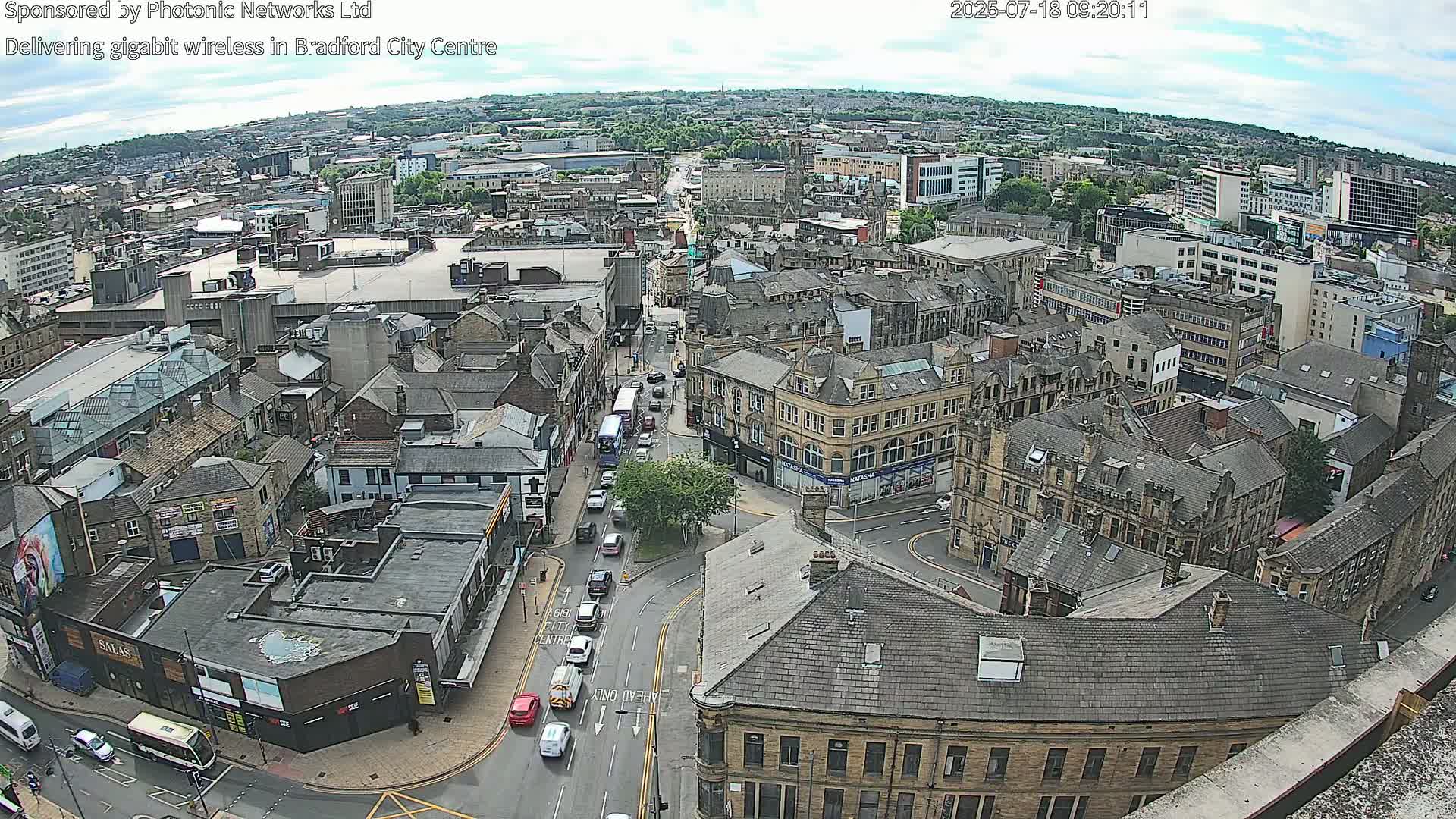 A high-angle, long shot of a city center on a partly cloudy day, showing many buildings, roads with cars, and some green spaces.