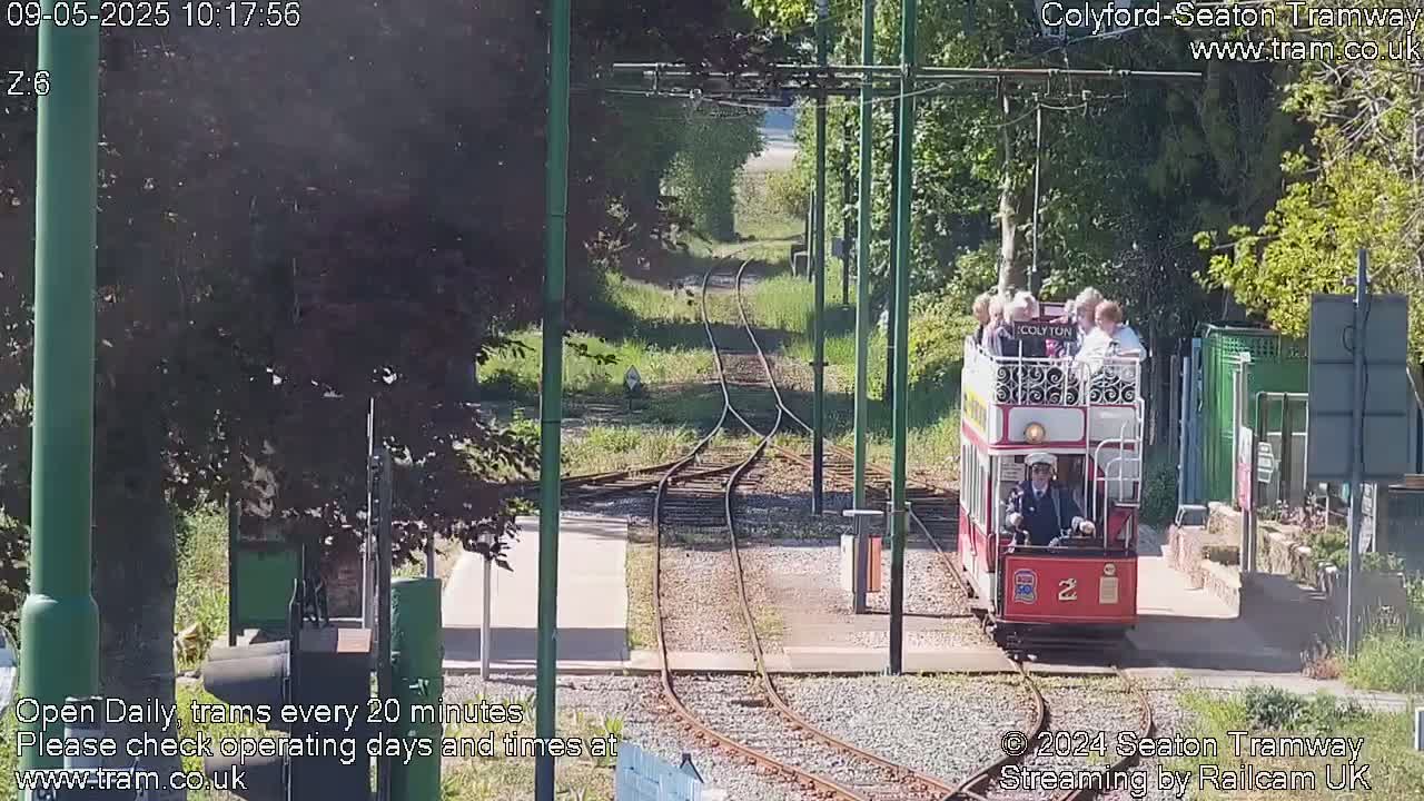 A red vintage-style tram carrying passengers approaches a station on a sunny day along a narrow gauge railway track lined with trees.