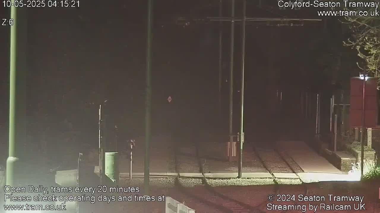 A nighttime view of a tram track with overhead wires, a small trash receptacle, and some vegetation visible in the dark.
