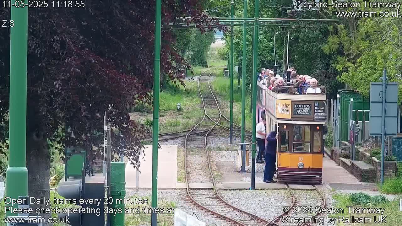 A vintage-style tram carrying passengers departs from a station on a sunny day, traveling along tram tracks surrounded by greenery.