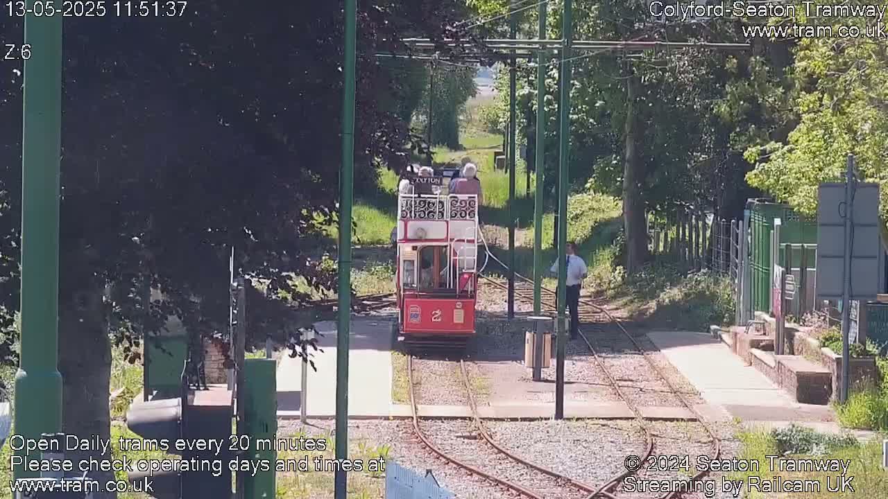 A red and white vintage tram is approaching a station on a sunny day, surrounded by green trees and foliage.
