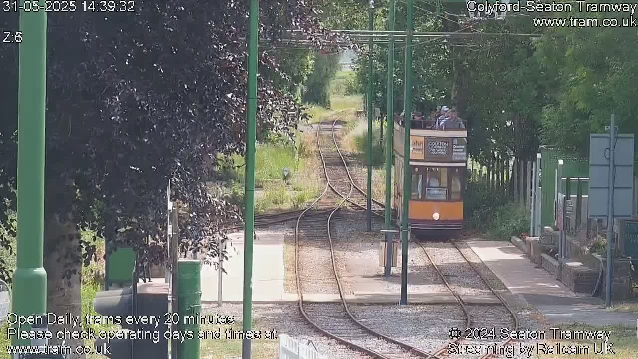 A yellow and brown vintage tram carrying passengers approaches a station on a sunny day.