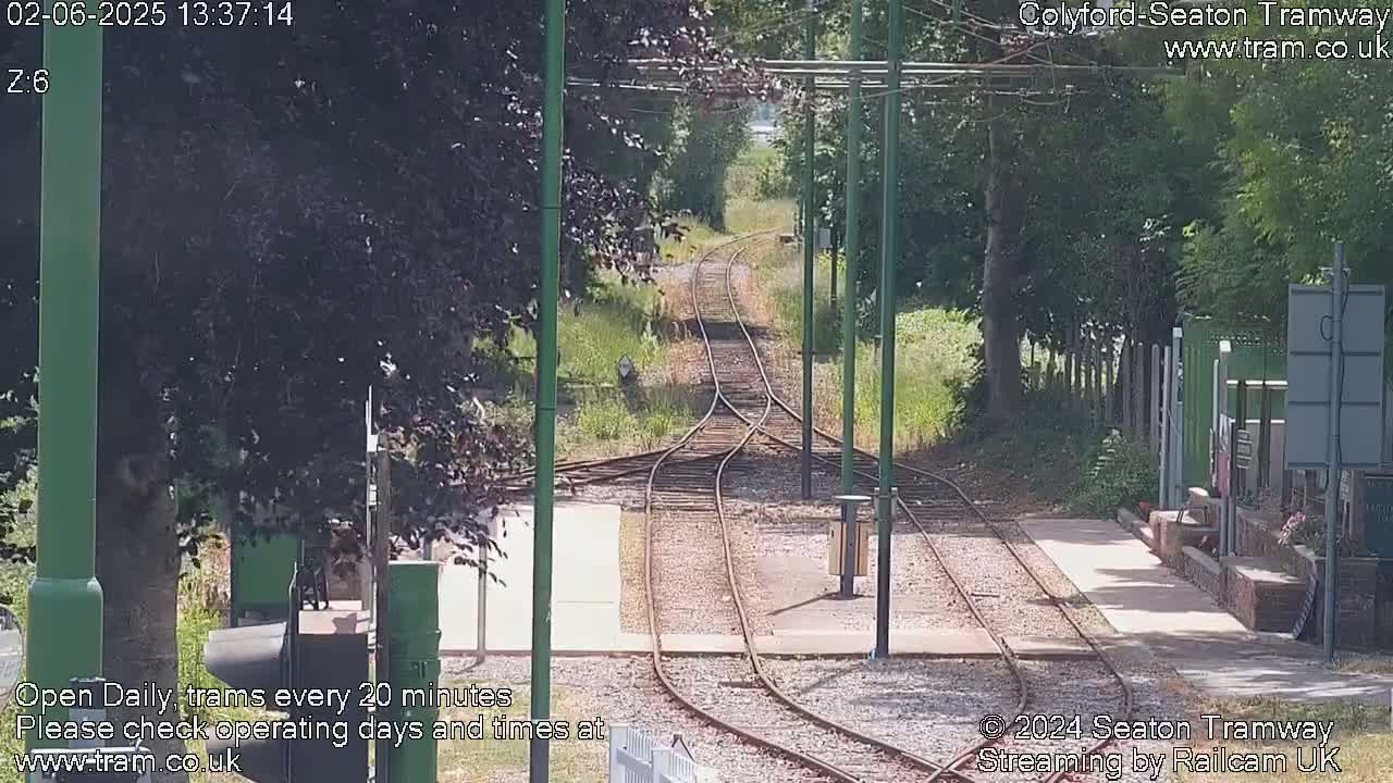 A sunny day reveals a split of tram tracks curving through a lush green landscape, bordered by trees and a paved platform.