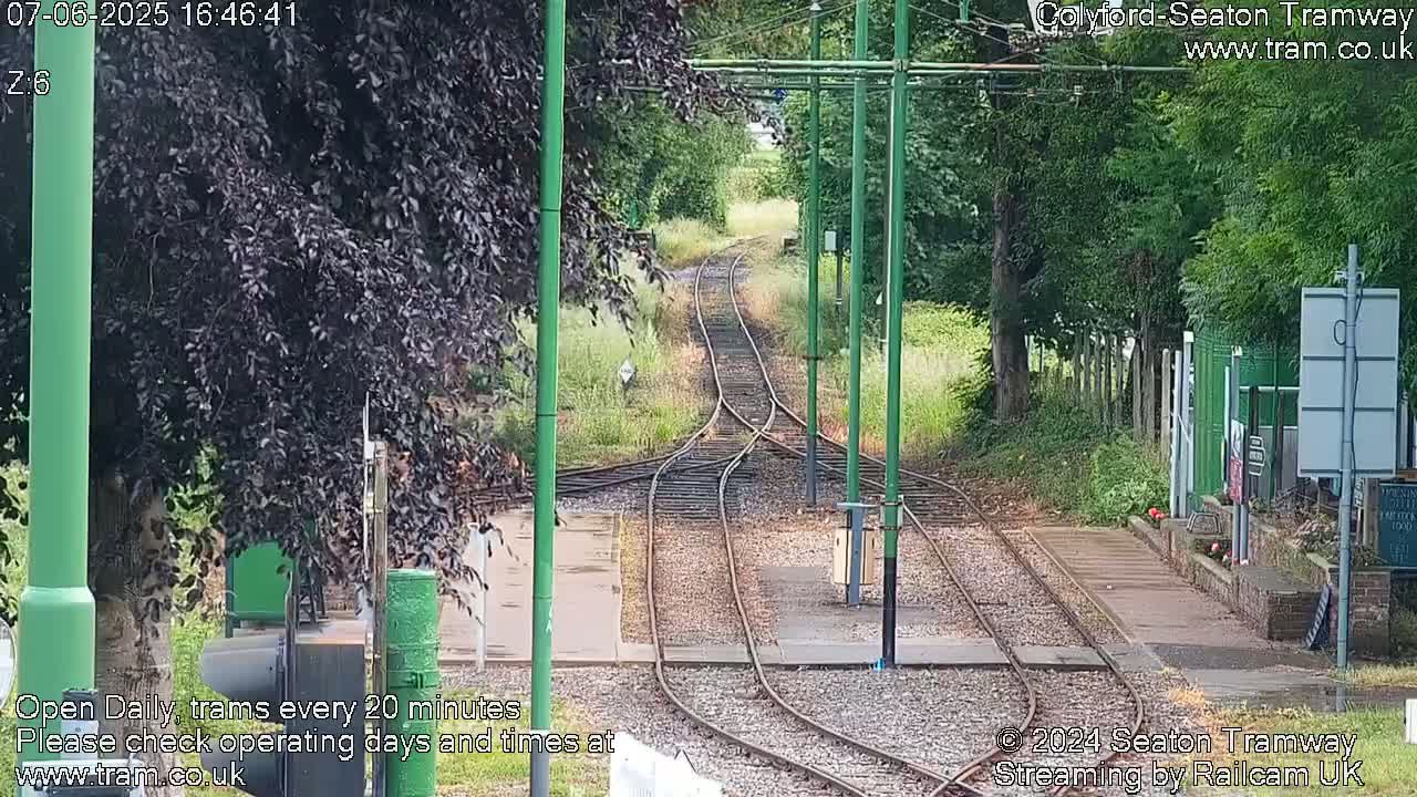 Tram tracks diverge at a point where green vegetation borders a paved walkway, under a partly cloudy sky.