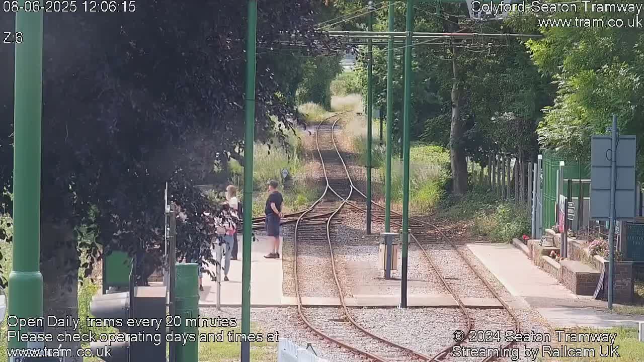A sunny outdoor scene shows a railway with three sets of tracks curving away from a platform where a few people stand.