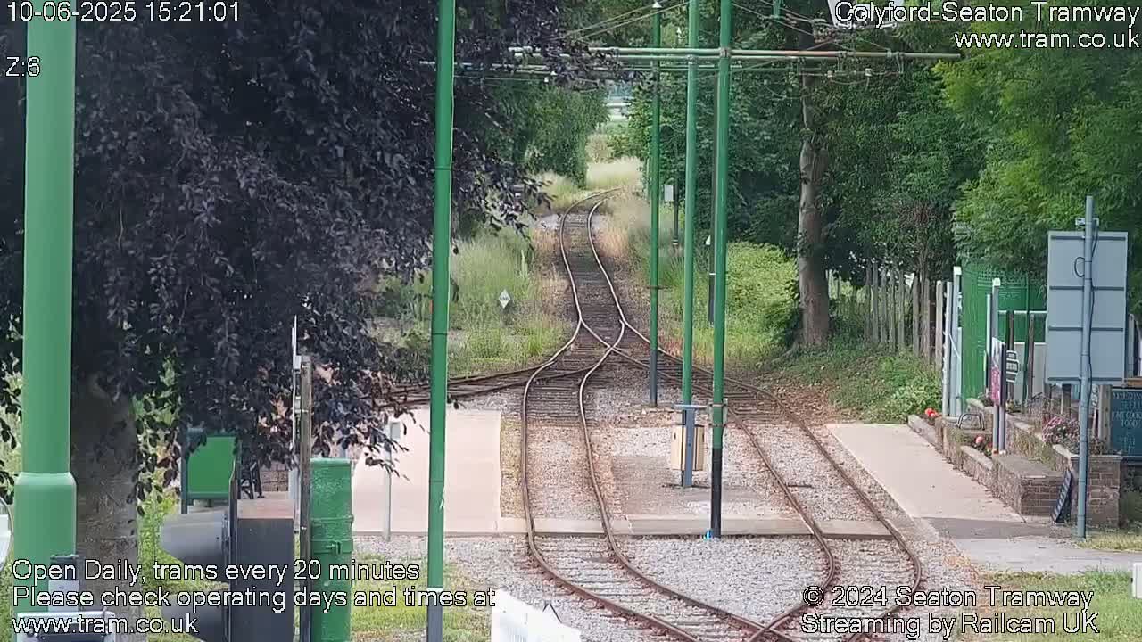 Tram tracks diverge in a grassy area bordered by trees under a partly sunny sky.