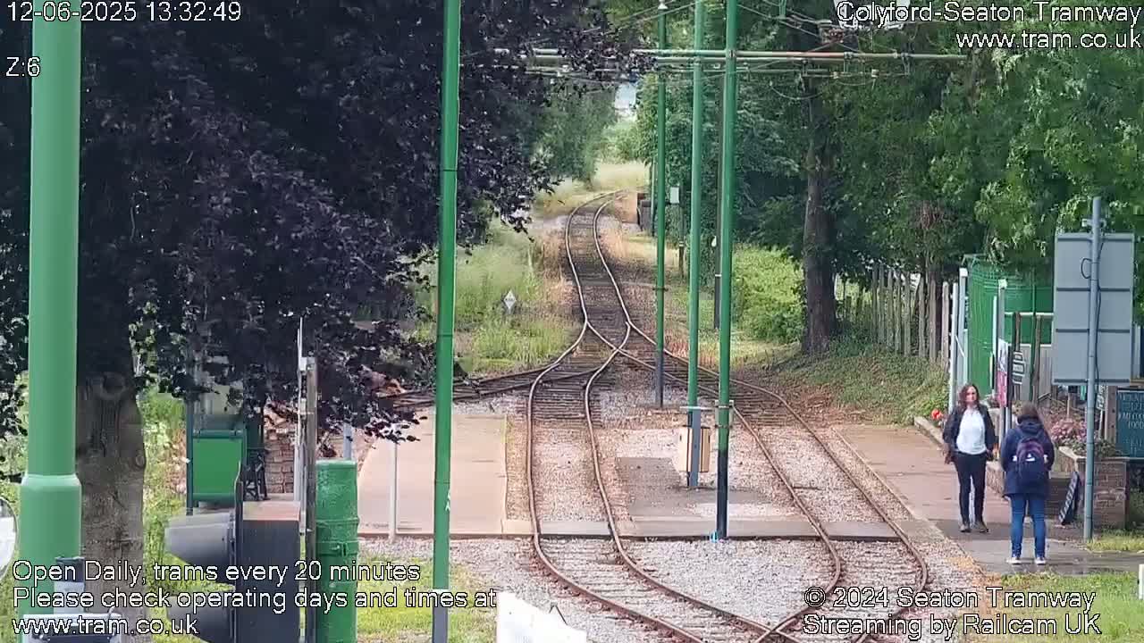 Two people stand near a tram track junction in a wooded area on a partly cloudy day.