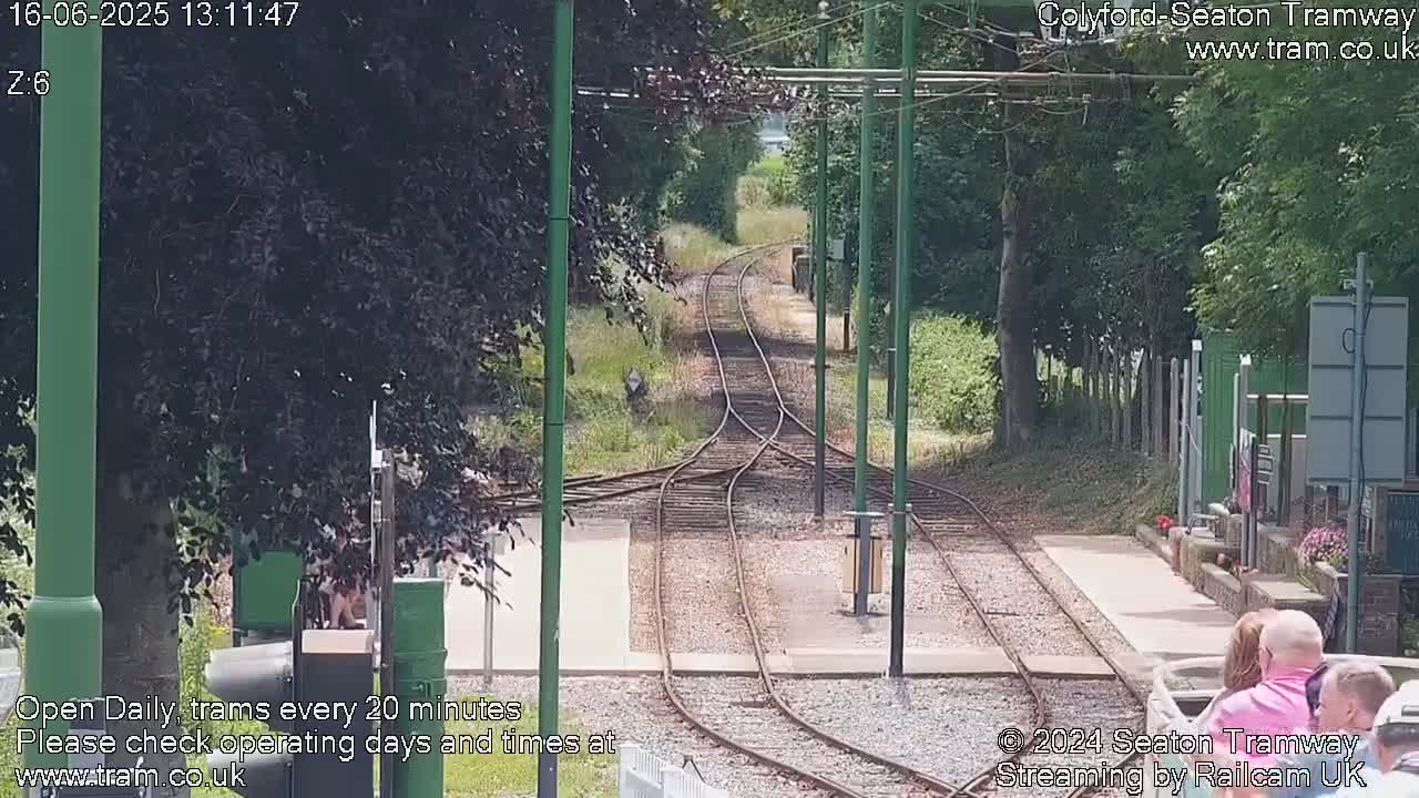 A sunny day at a tram station shows a split of tram tracks leading into a tree-lined area, with several people standing nearby.