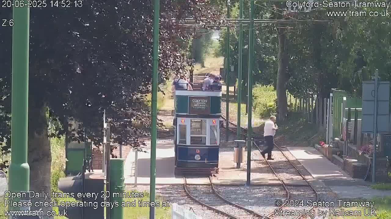 A vintage-style tram carrying passengers approaches a station on a sunny day, surrounded by lush green trees.