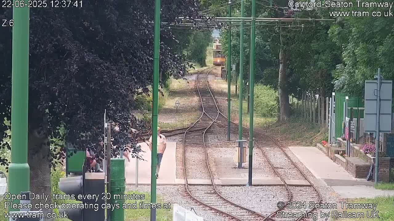 A yellow tram travels down a track toward a station on a sunny day, with people standing near the tracks and trees lining the path.