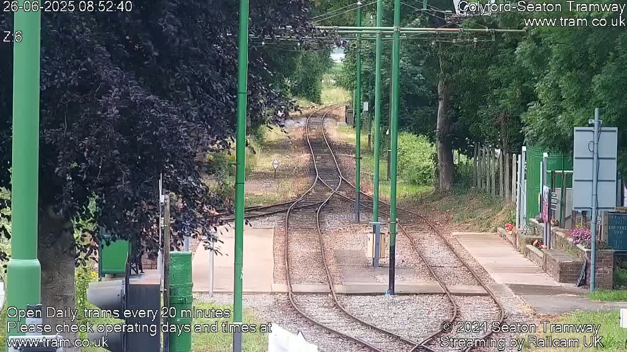 A view of train tracks splitting into two directions at a station on a sunny day, surrounded by lush green trees and vegetation.