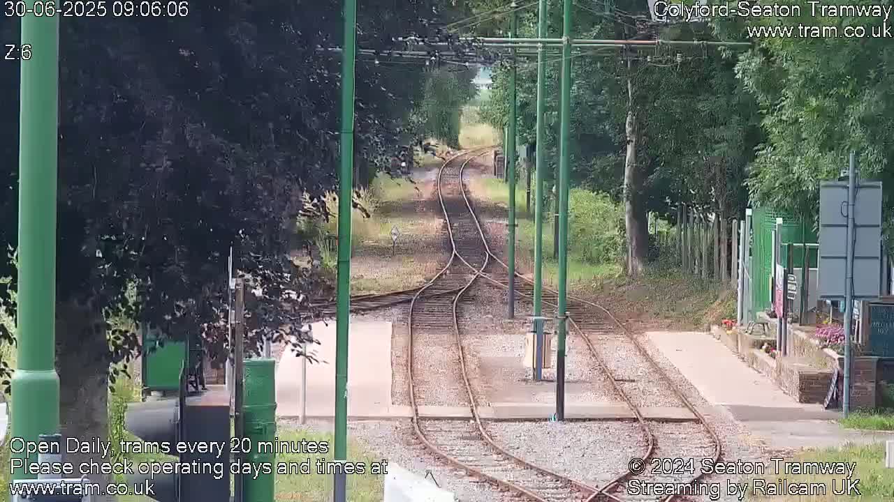 A view of train tracks splitting into two directions, bordered by lush green trees and shrubs on a partly sunny day.